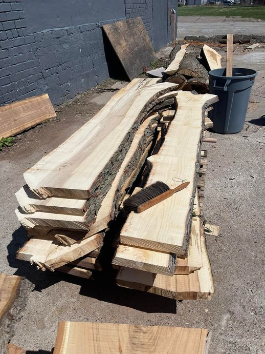 Stacked rough-cut lumber boards on gravel beside a building wall and trash bin outdoors.