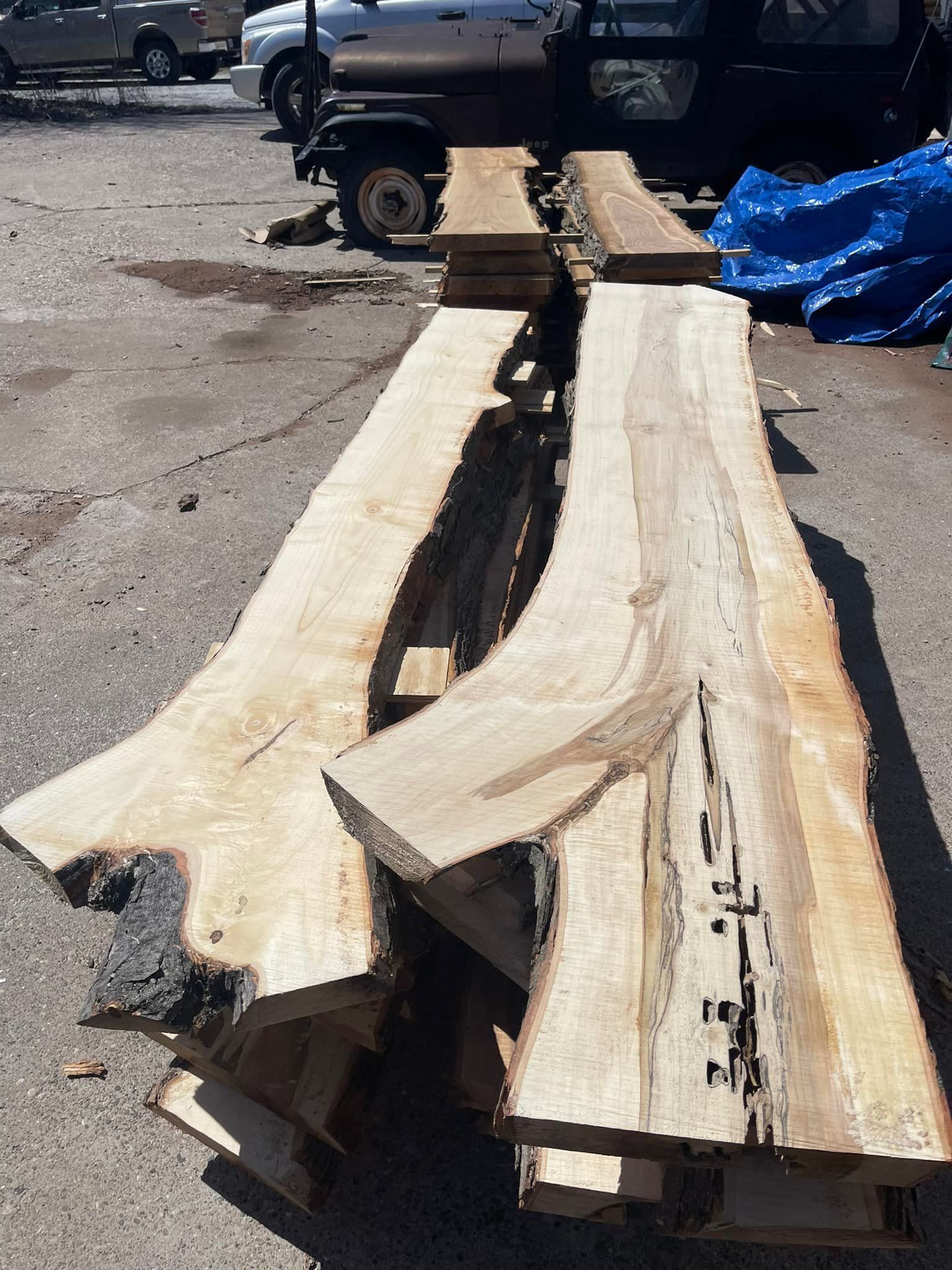 Rough-cut wooden planks stacked on a curb beside parked trucks, with a blue tarp in the background.