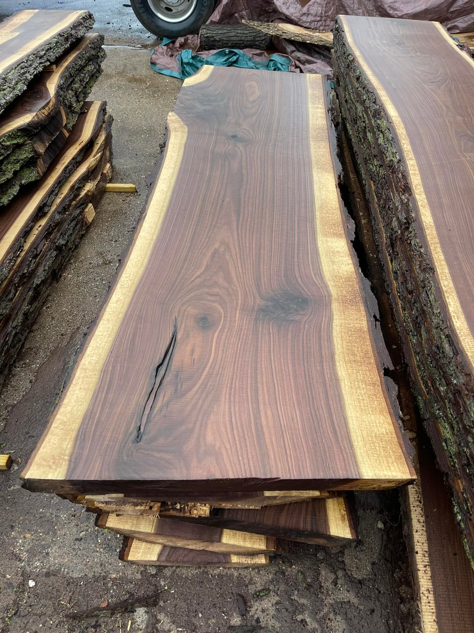 Stacked live-edge wooden slabs with dark grain and bark edges at an outdoor lumber yard.