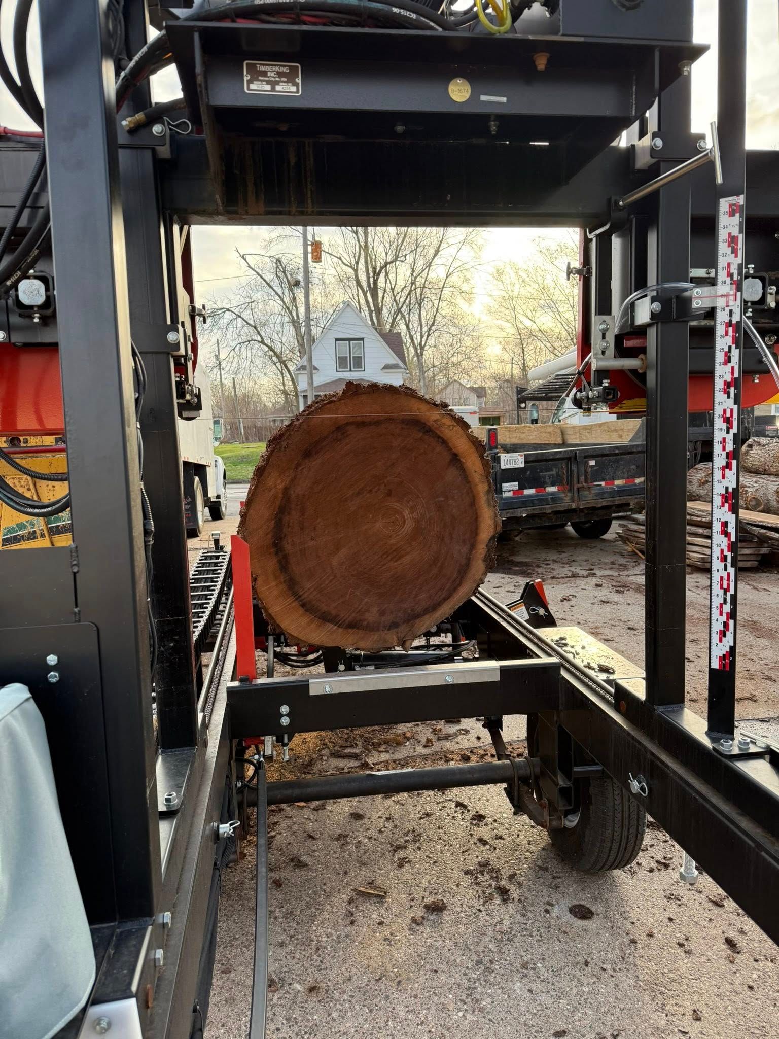 Large log in a wood splitter outdoors, with gravel ground and trees in the background