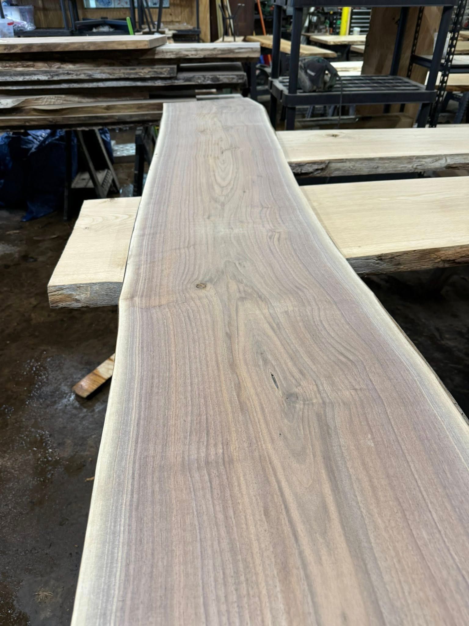 Long wooden plank resting on supports in a workshop, with rough-cut boards stacked around it.