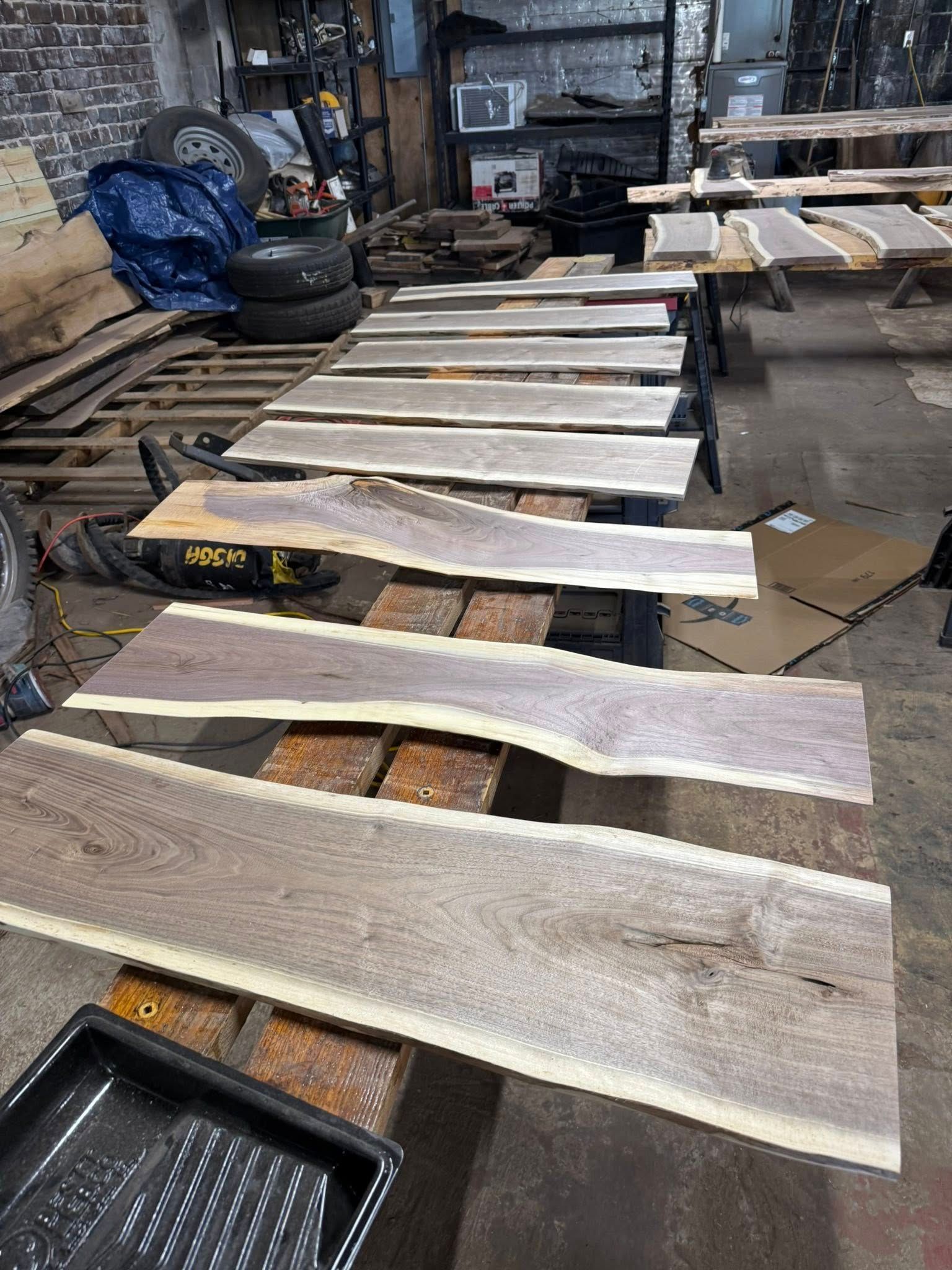 Long wooden boards laid out on workbenches in a cluttered workshop, with tools and materials nearby