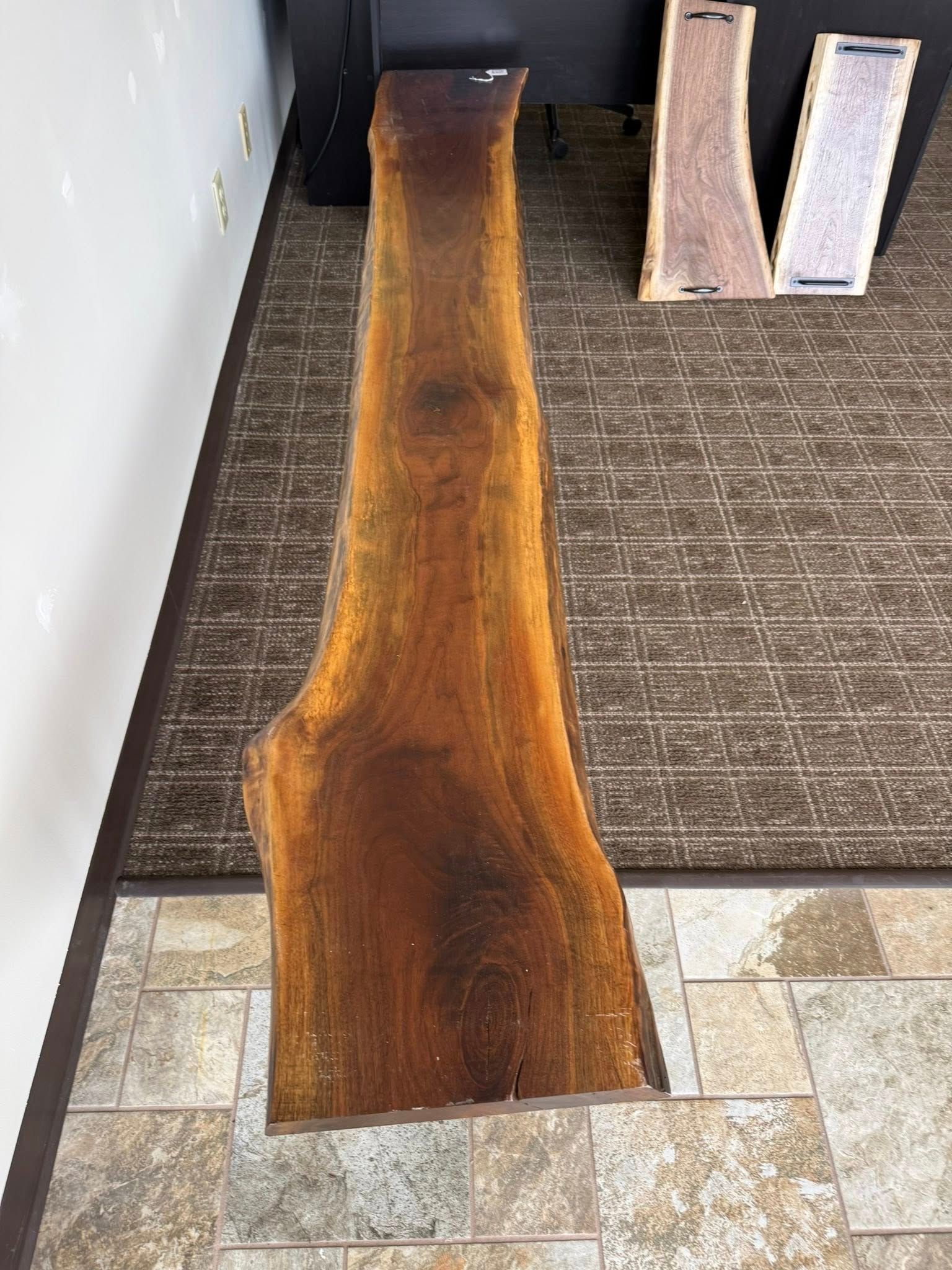 Wooden plank with a natural live-edge grain, laid on a carpeted floor beside a wall and foam blocks.