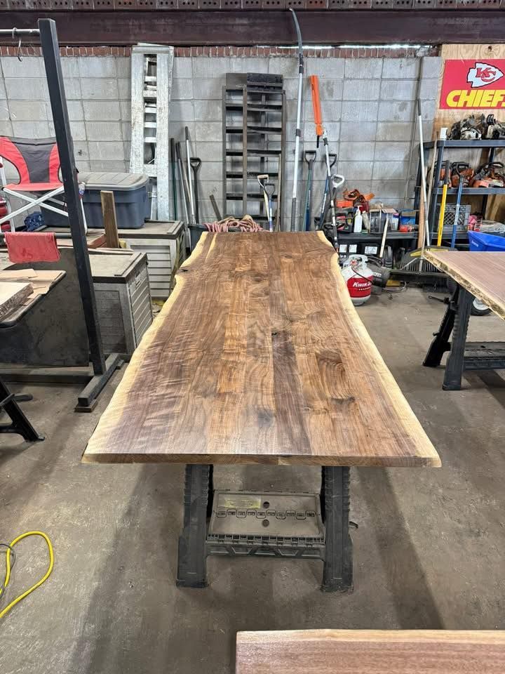 Long wooden tabletop on metal stands in a workshop, with shelves and tools in the background.