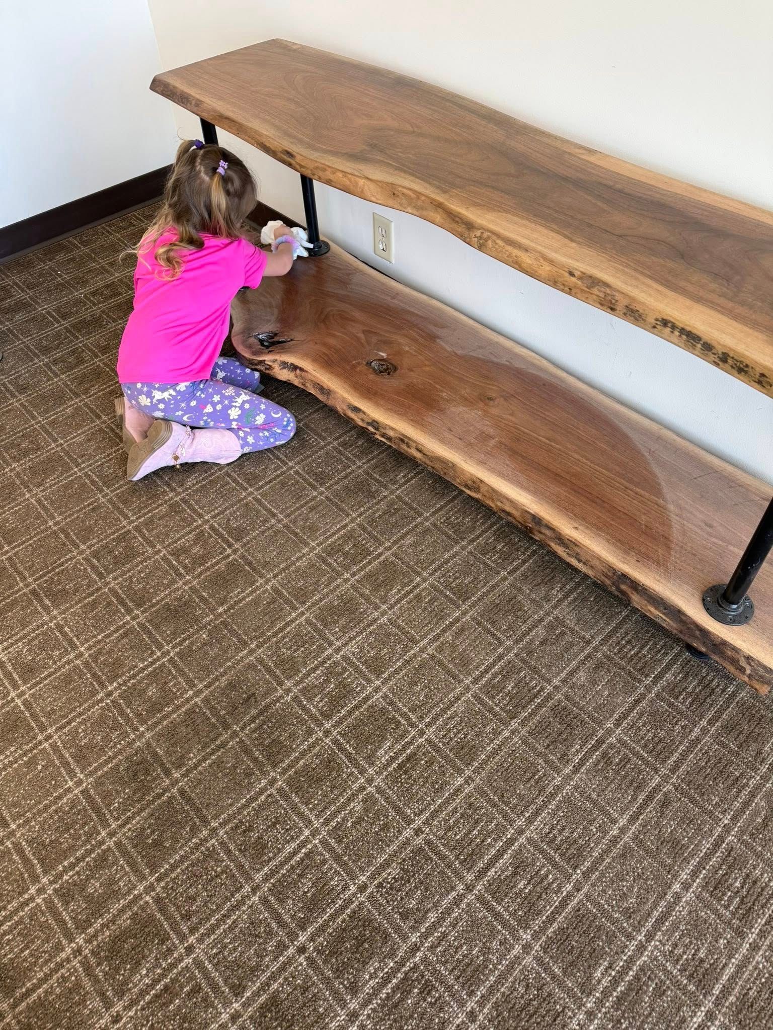Child in pink shirt kneels at a wooden bench indoors, reaching toward the lower shelf.
