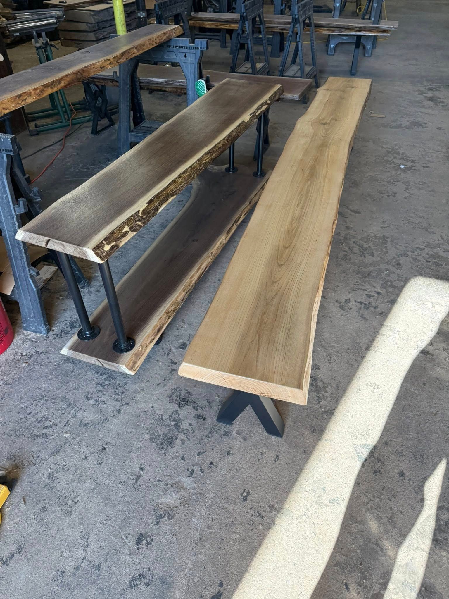 Long narrow wooden planks on metal stands in a workshop, with sawdust on the concrete floor.