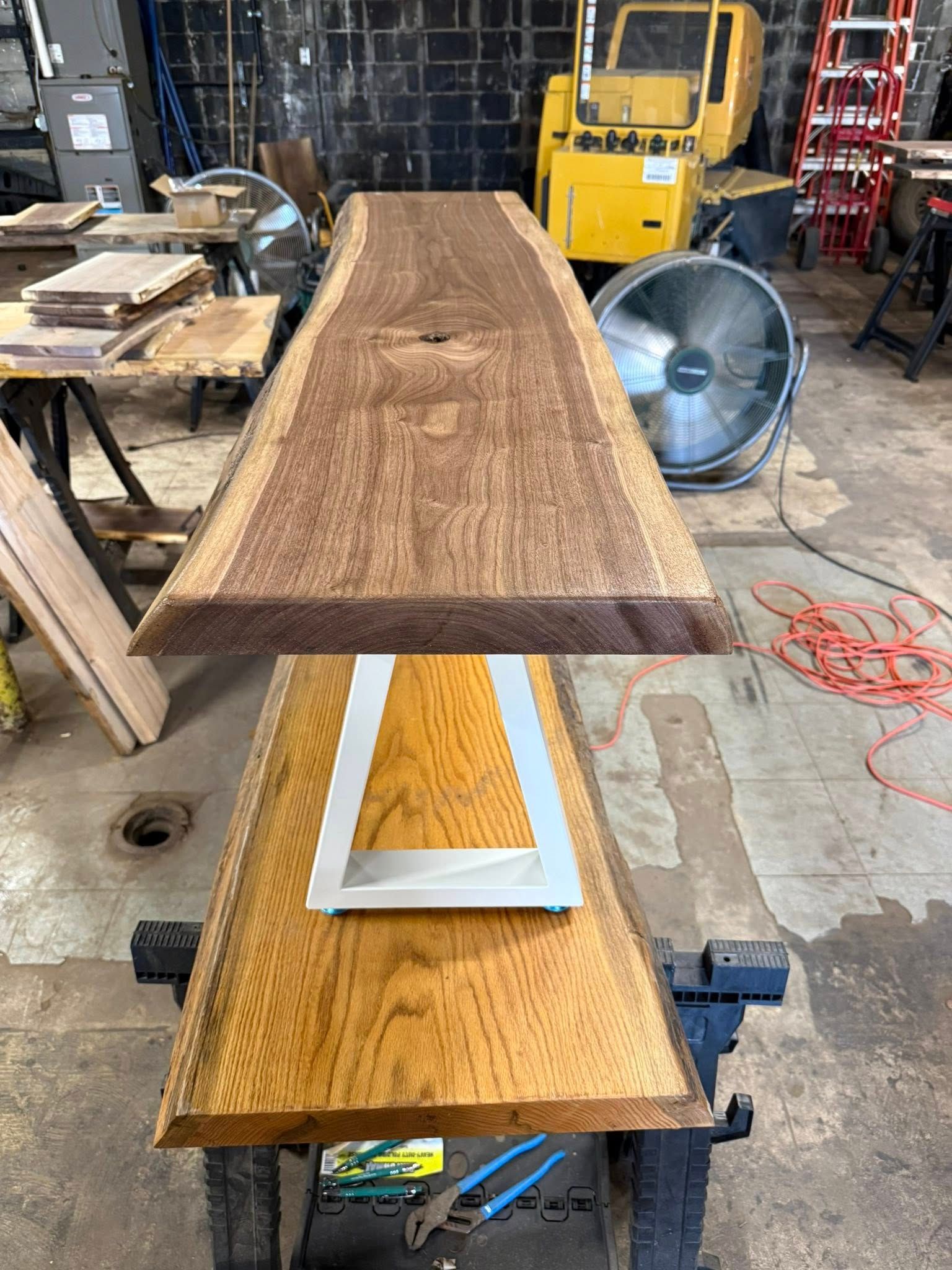 Two stacked wooden slabs on a metal stand in a workshop, with a fan and tools in the background.