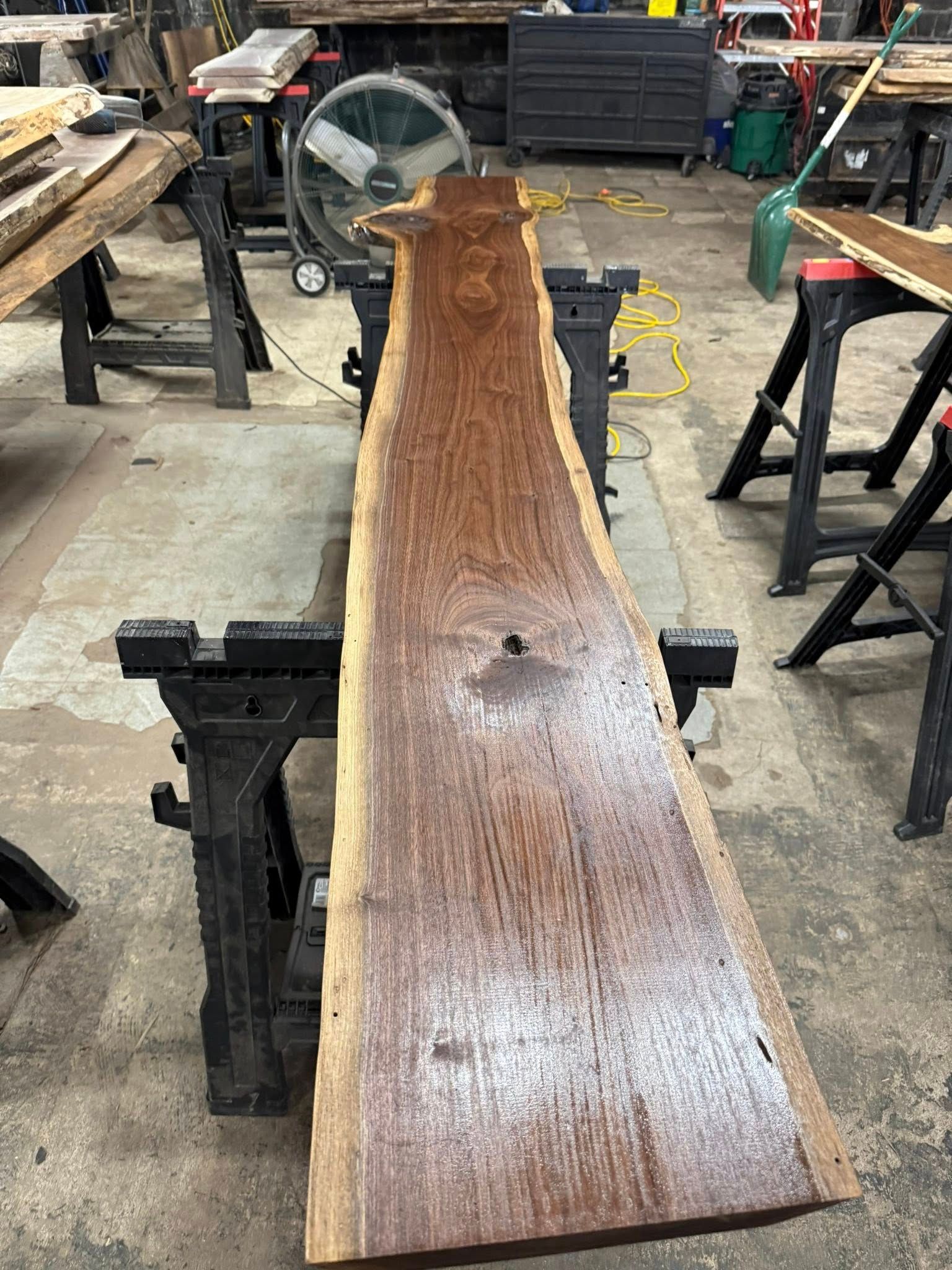 Long, narrow live-edge wood slab on workshop sawhorses, with a fan and tools in the background.