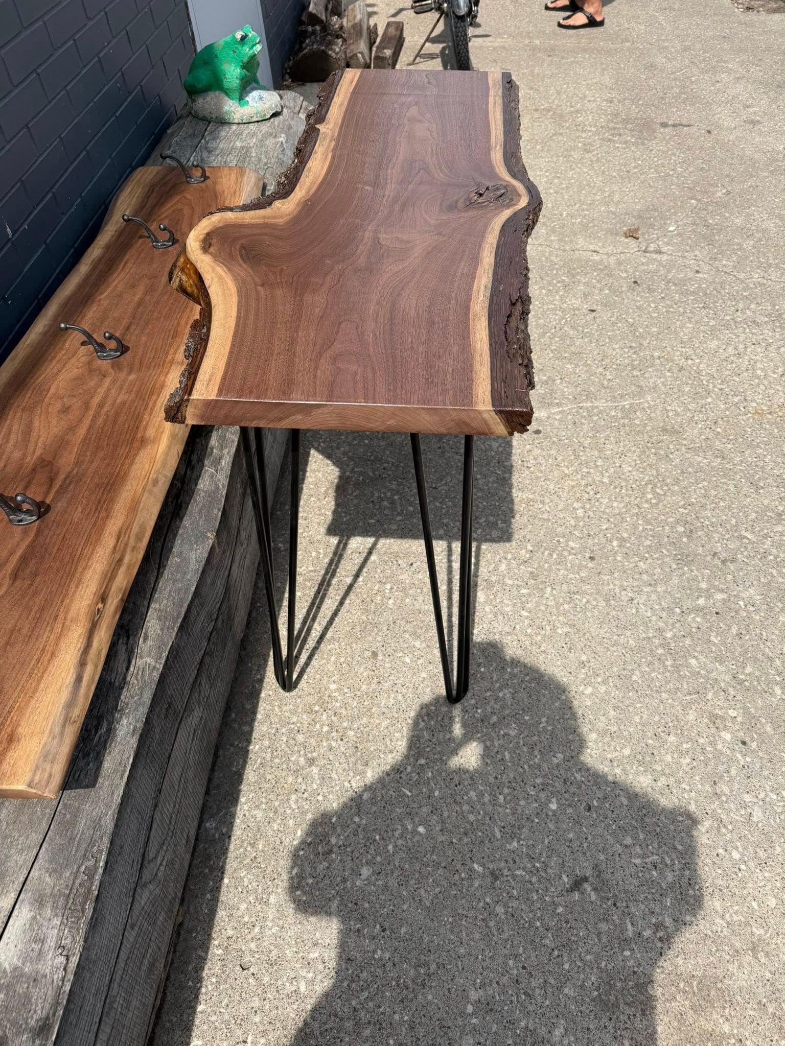 Wooden live-edge table top on metal hairpin legs displayed outdoors on pavement and a bench.