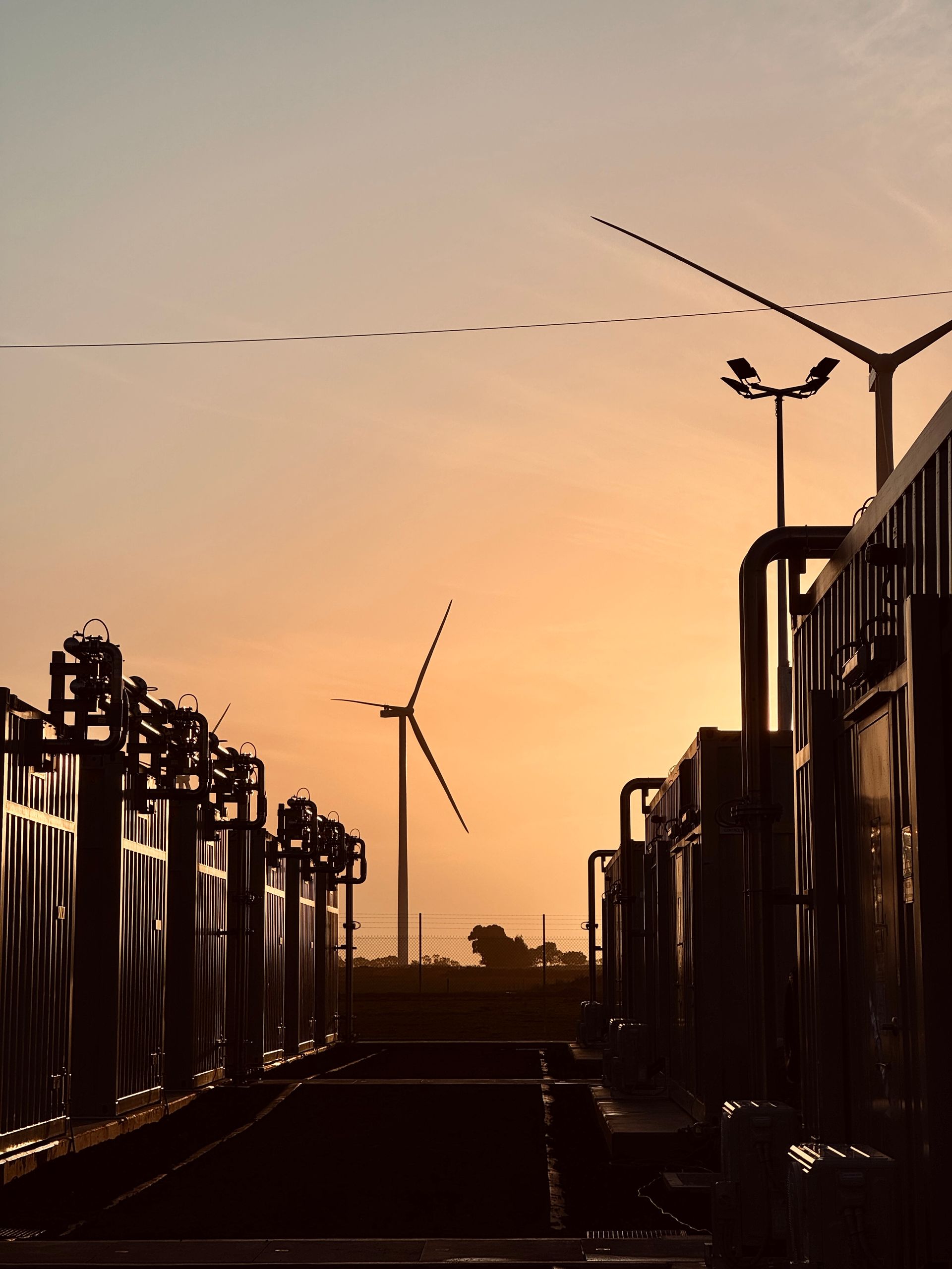 Sunset over wind turbine and industrial equipment. Silhouetted structures against orange sky.