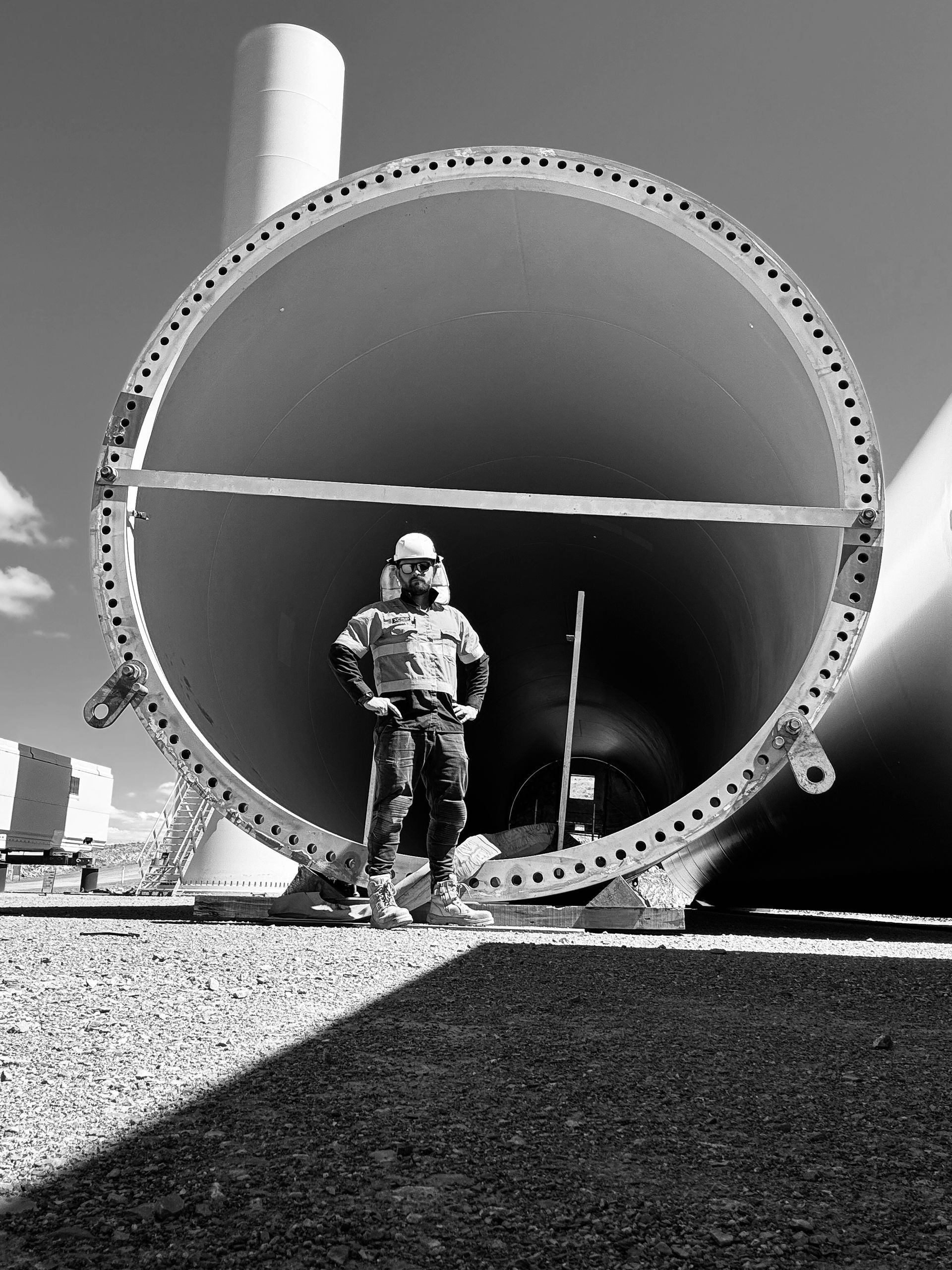 Worker stands inside large wind turbine component, wearing safety gear, with the sky and other parts in the background.