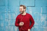 Man wearing red sweater, listening to music on earbuds, smiling, holding phone, standing against blue brick wall.