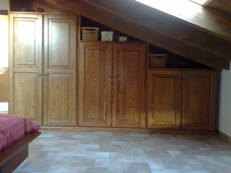 Armadietti in legno ricavati in una soffitta con soffitto spiovente e pavimento in piastrelle quadrate.