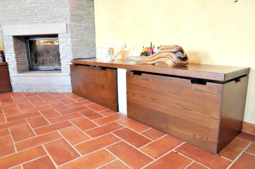 Mobile contenitore in legno con parte superiore aperta, contro una parete gialla e un pavimento in mattoni.