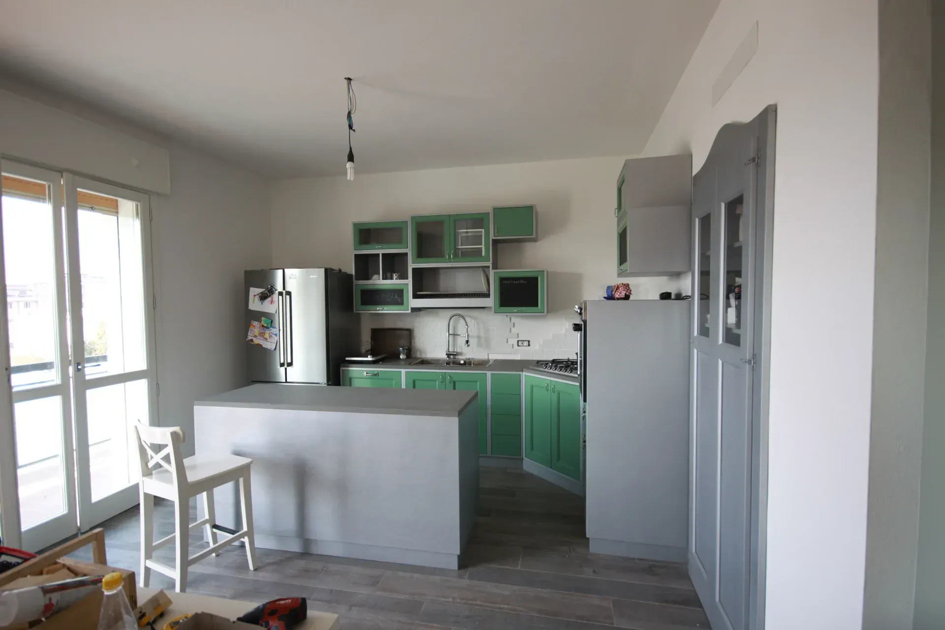 Cucina con mobili verdi, isola grigia, elettrodomestici in acciaio inossidabile e uno sgabello da bar bianco.