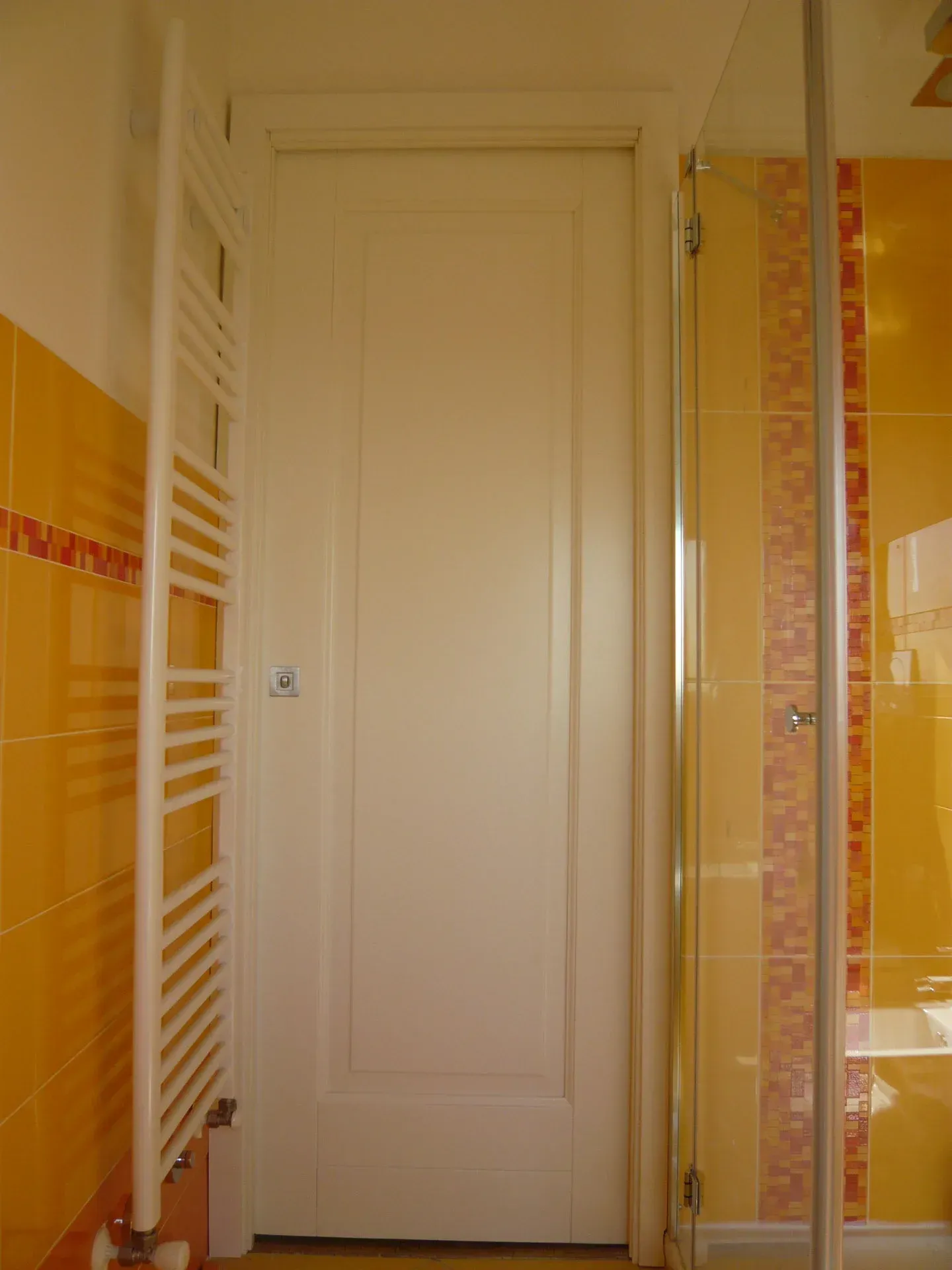 Porta bianca del bagno tra un termosifone bianco e una doccia in vetro, in contrasto con pareti piastrellate gialle e rosse.