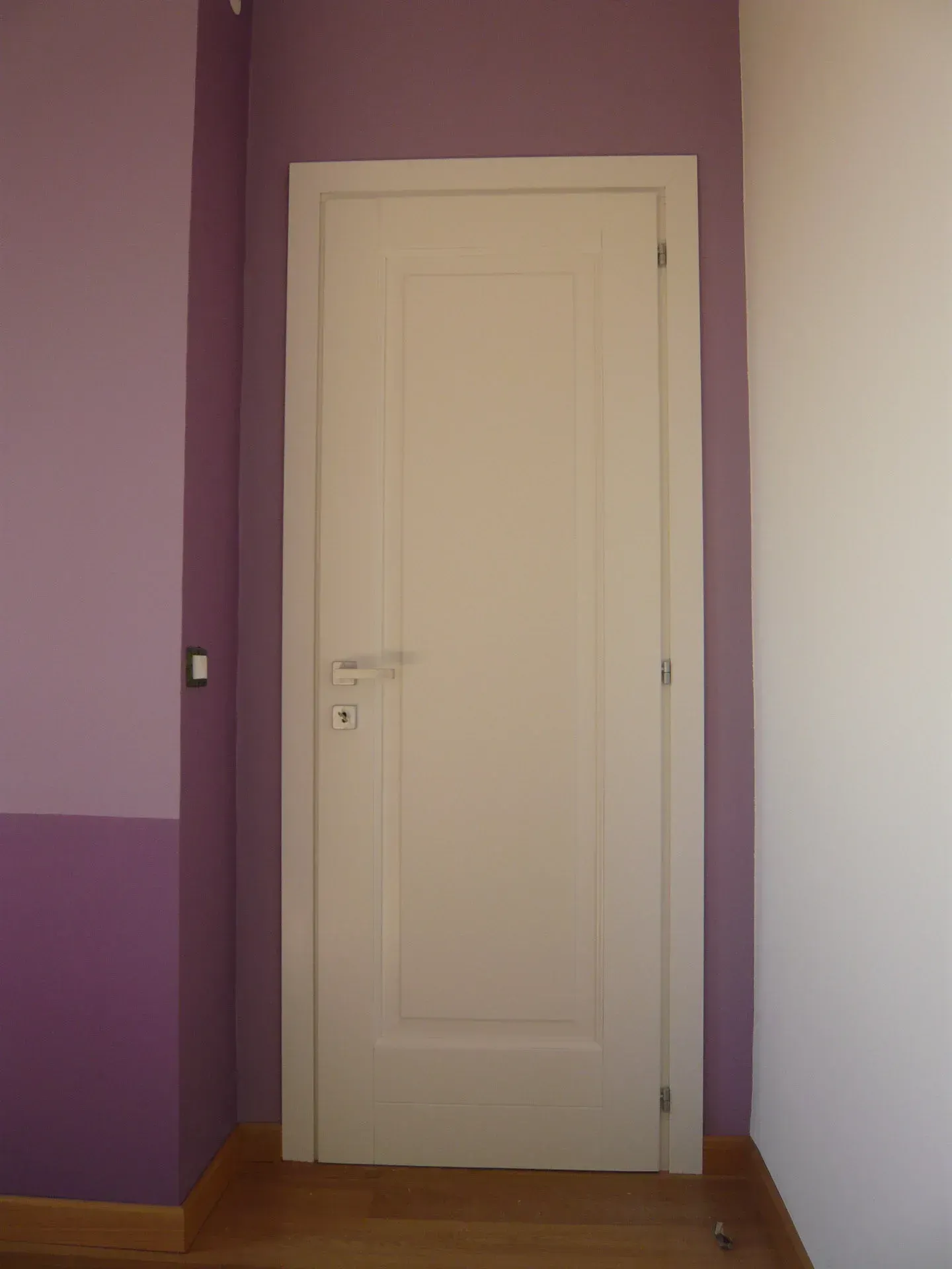 Porta bianca incastonata in una parete dipinta di viola e bianco; pavimento in legno.