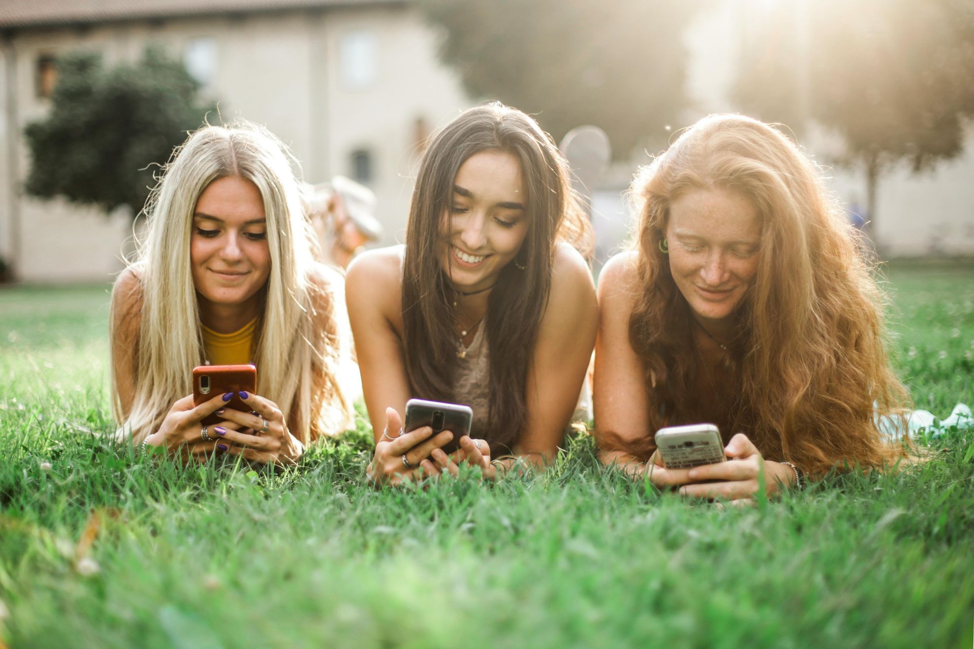 Three young women lying on grass, smiling while looking at their phones.