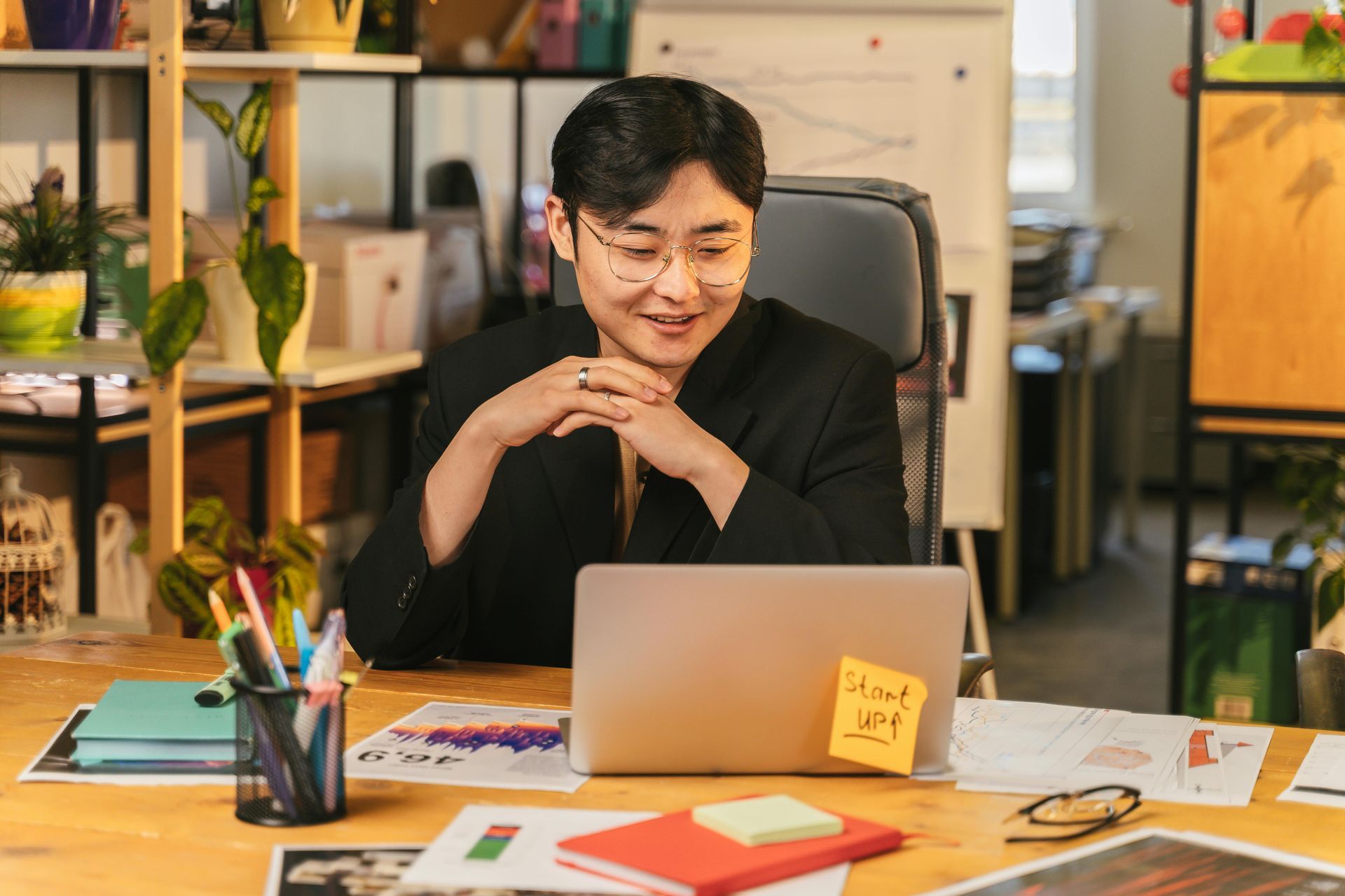 Person in glasses at a desk looking at a laptop; office setting with plants and notes.