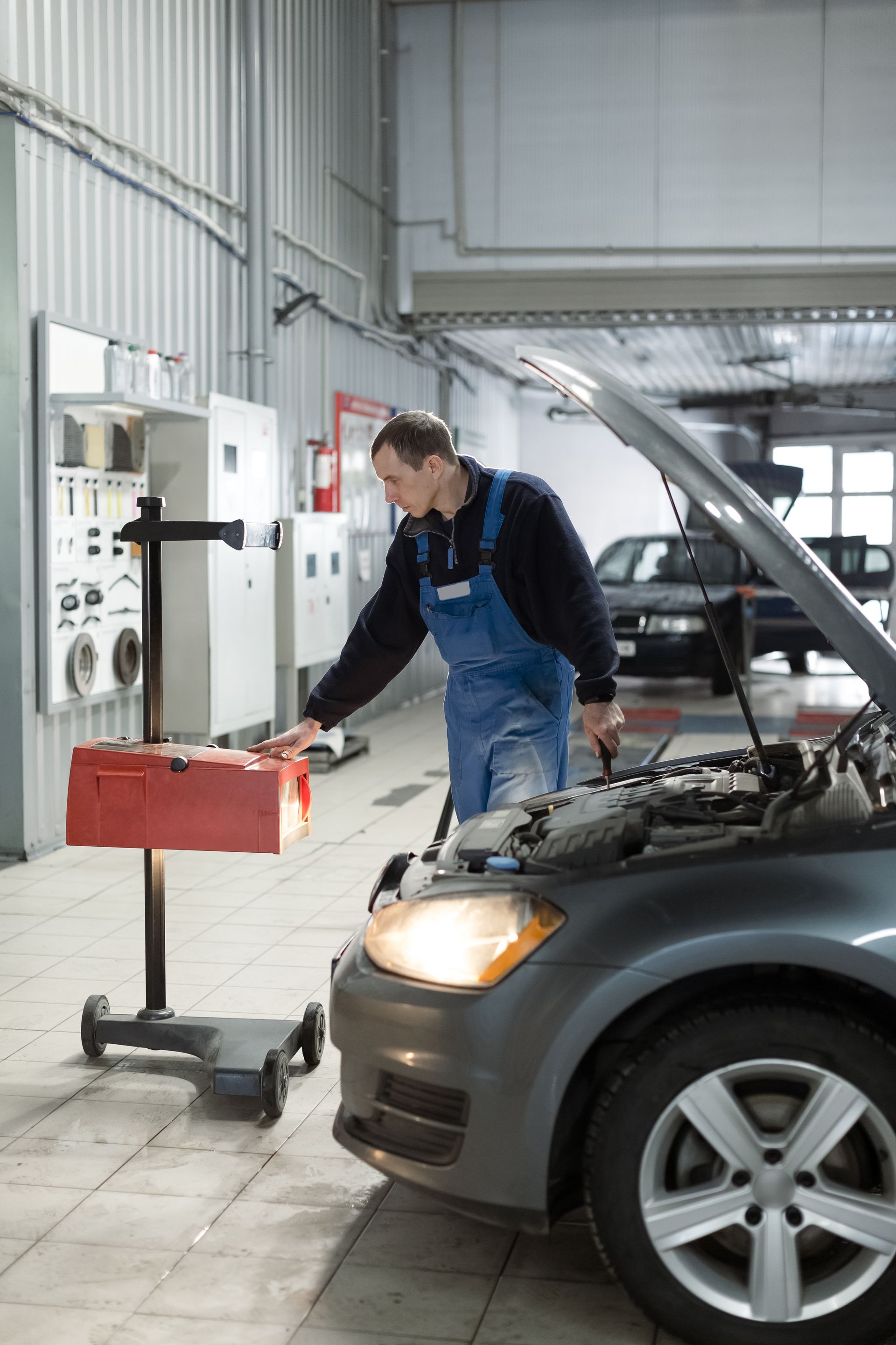 Mechanic in blue overalls checking car headlight in garage with a testing device.
