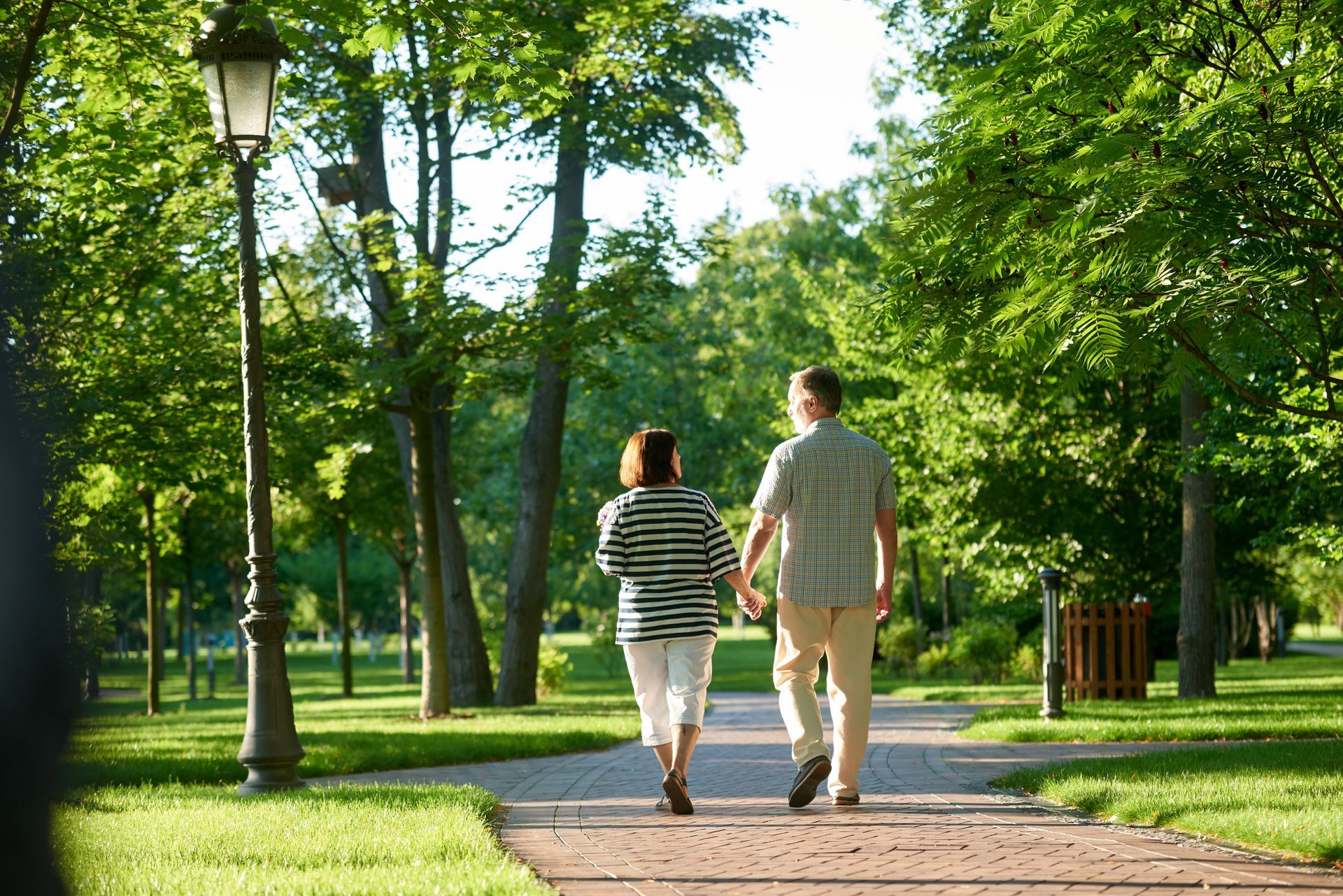 Couple walking hand-in-hand on a brick path in a sunny park, trees and a lamppost on either side.