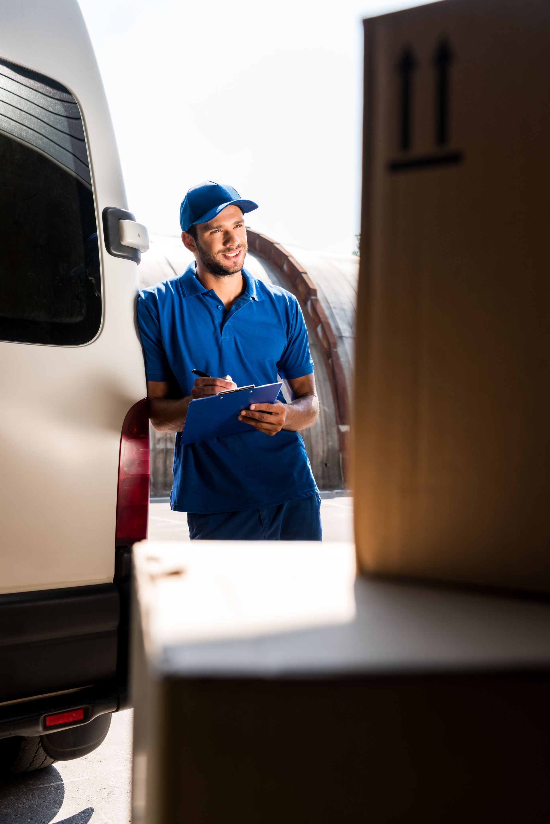 Delivery person in blue uniform leans against van, writing on a clipboard, boxes in foreground.