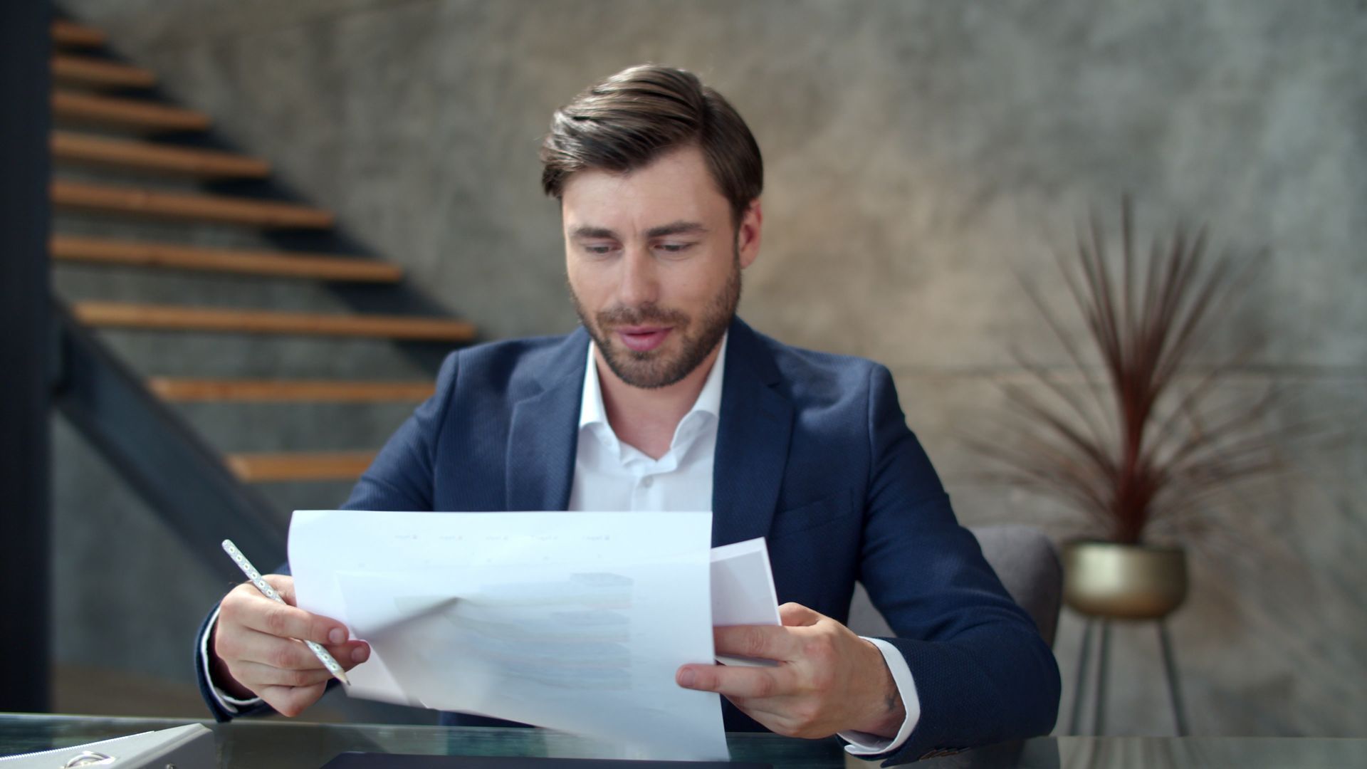 Man in blue suit, looking at documents, holding a pen. Inside, near stairs and a plant.