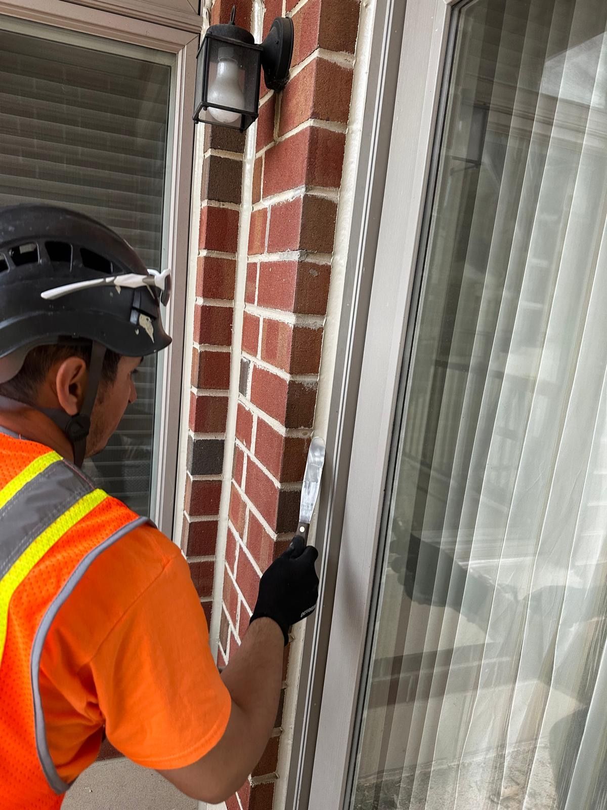 Person wearing safety gear applying sealant to brick wall beside a window.