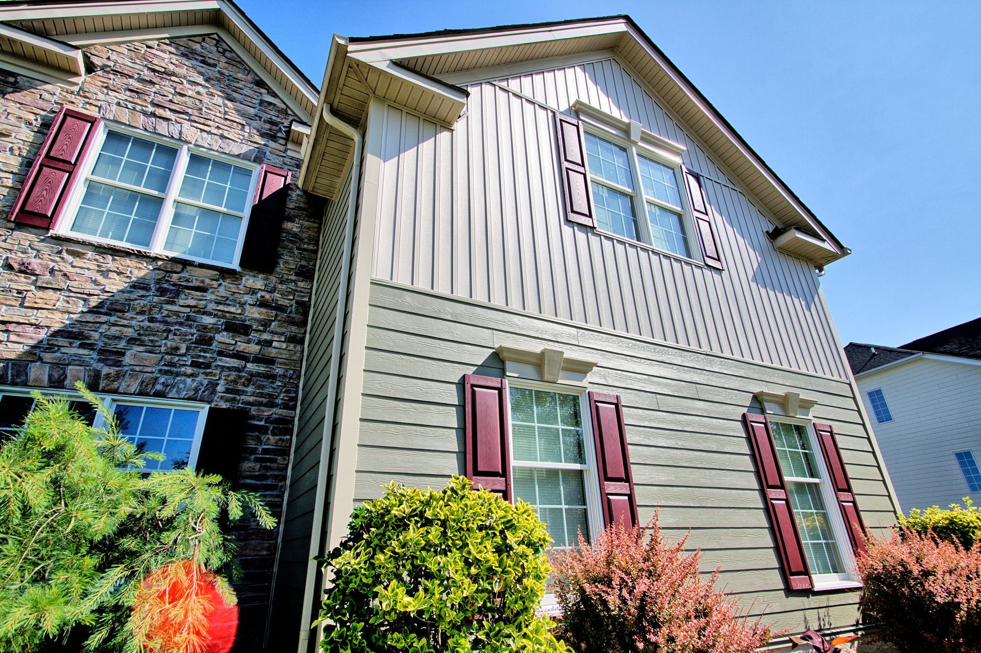 Two-story house with stone and gray siding, burgundy shutters, and a sunny blue sky.