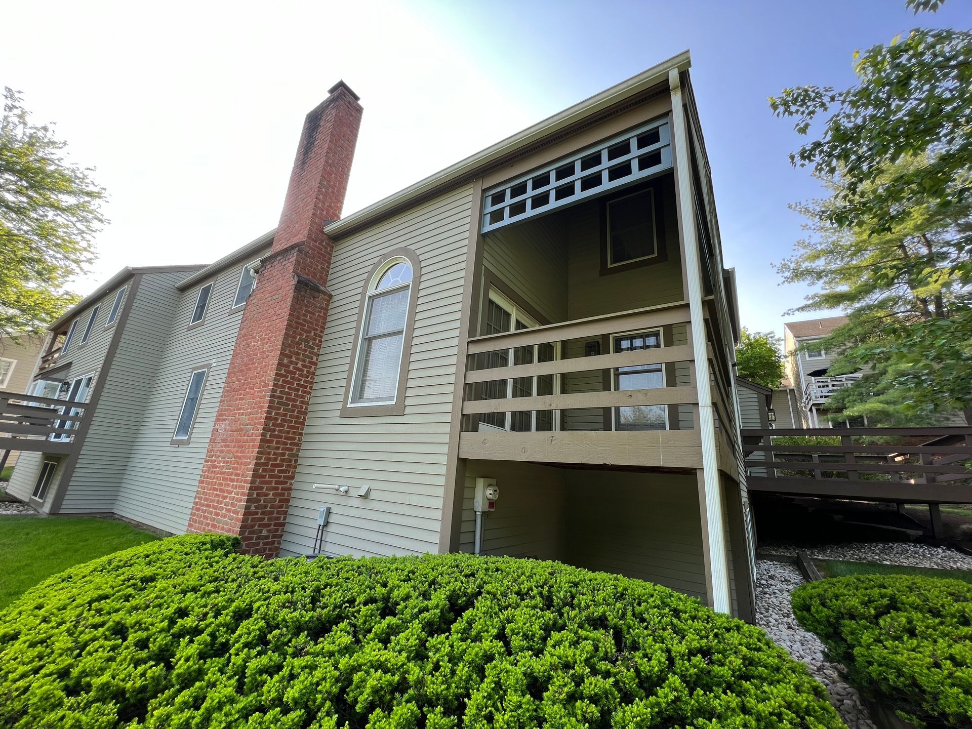 Beige two-story building with a brick chimney and wooden balcony, set in a green landscape with a footbridge.