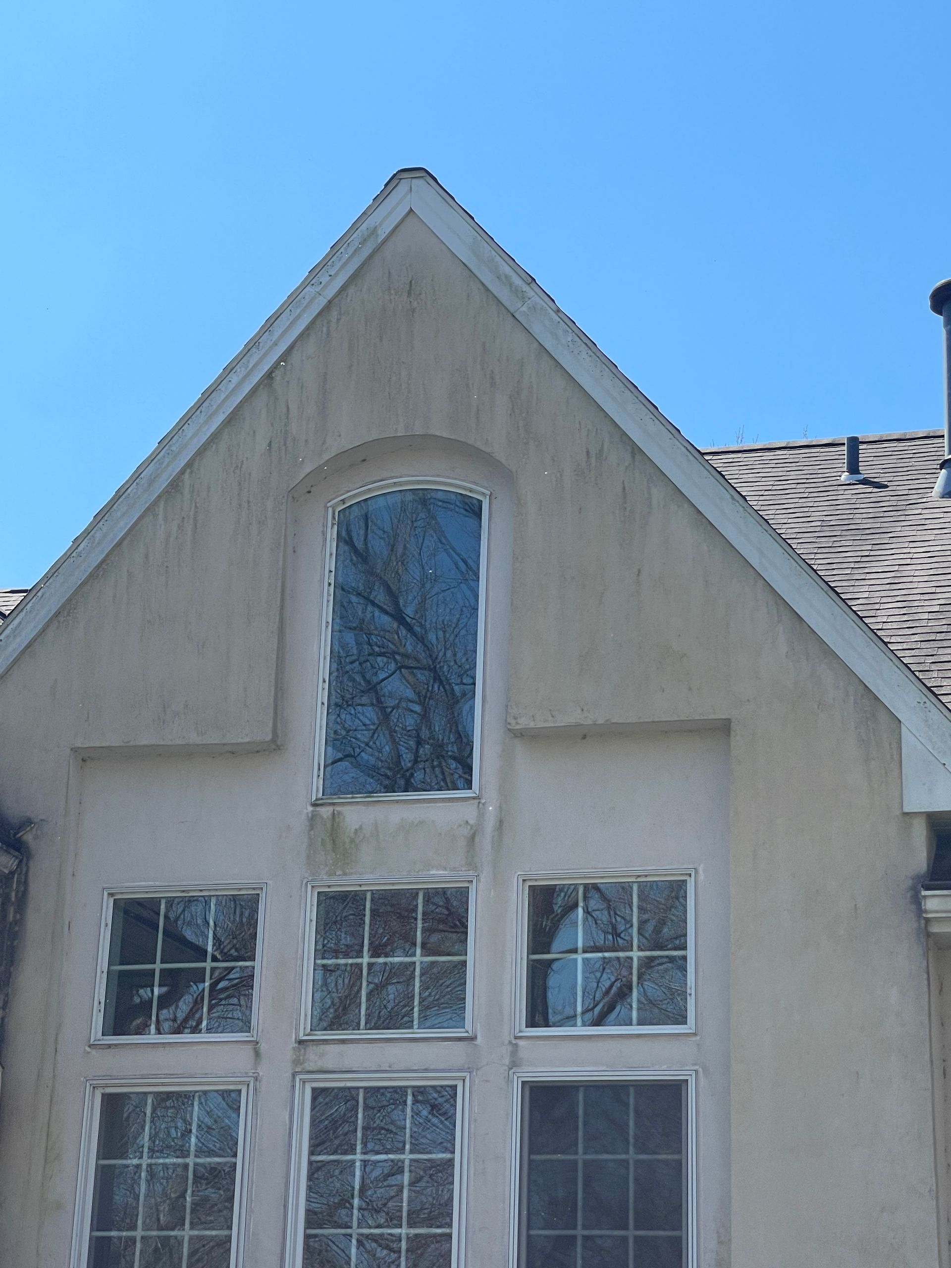 Beige house facade with six windows, an arched window, and a pointed roof against a blue sky.