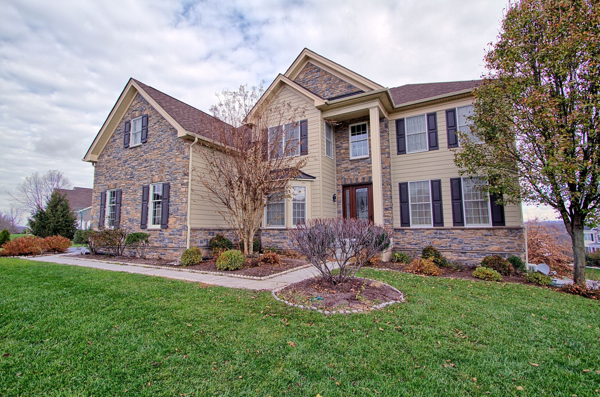 Two-story beige house with stone accents, black shutters, and a well-manicured lawn.