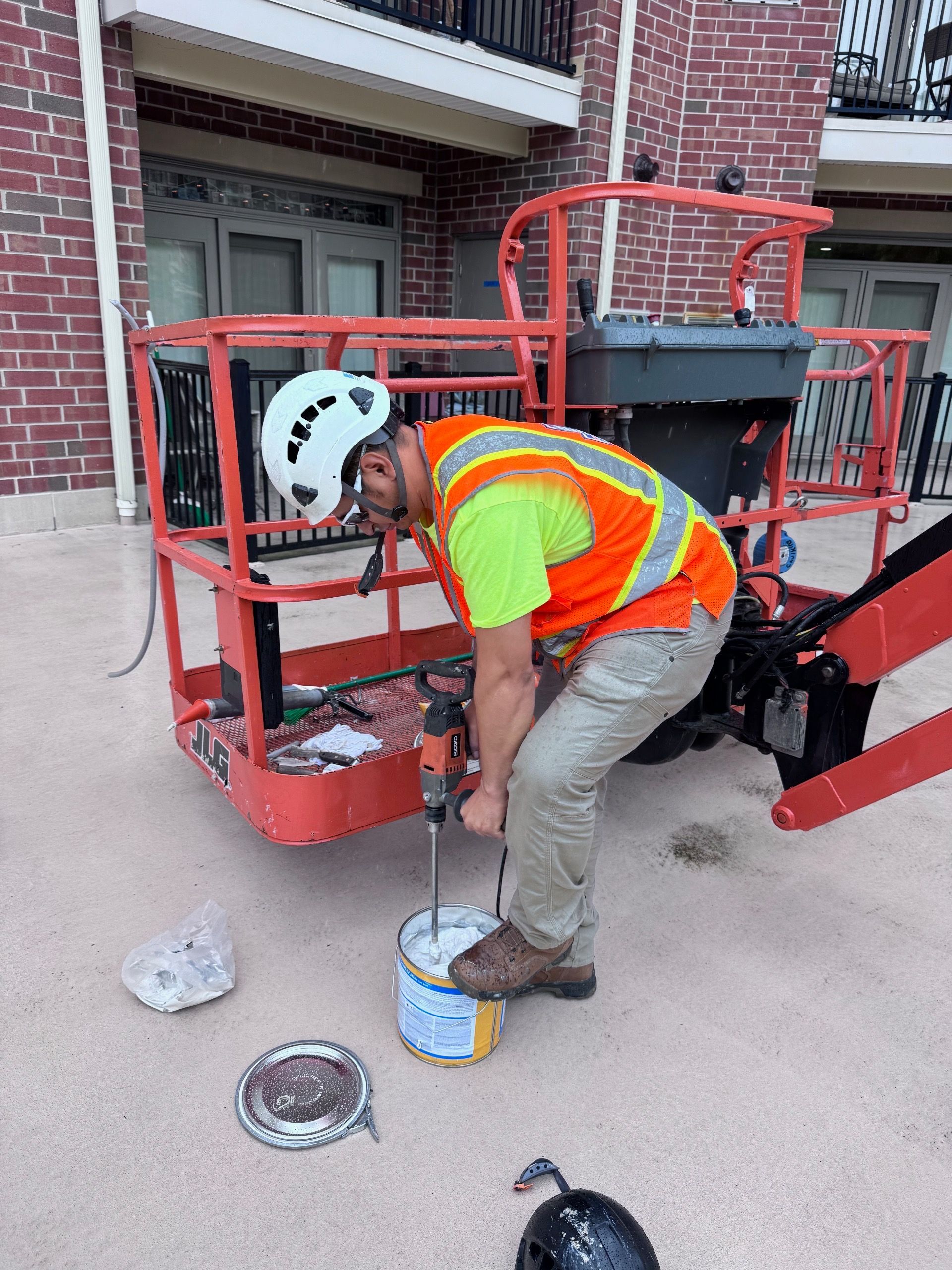 Construction worker mixing paint in a bucket while standing in a lift. He wears a hard hat and safety vest.