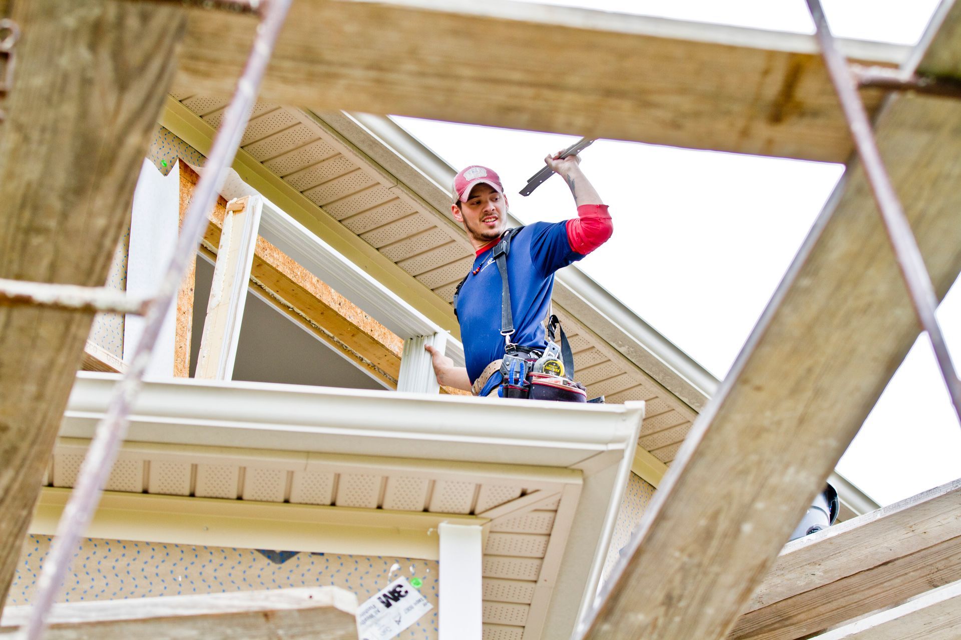 Man working on exterior trim, holding a tool, on a roof with a safety harness.