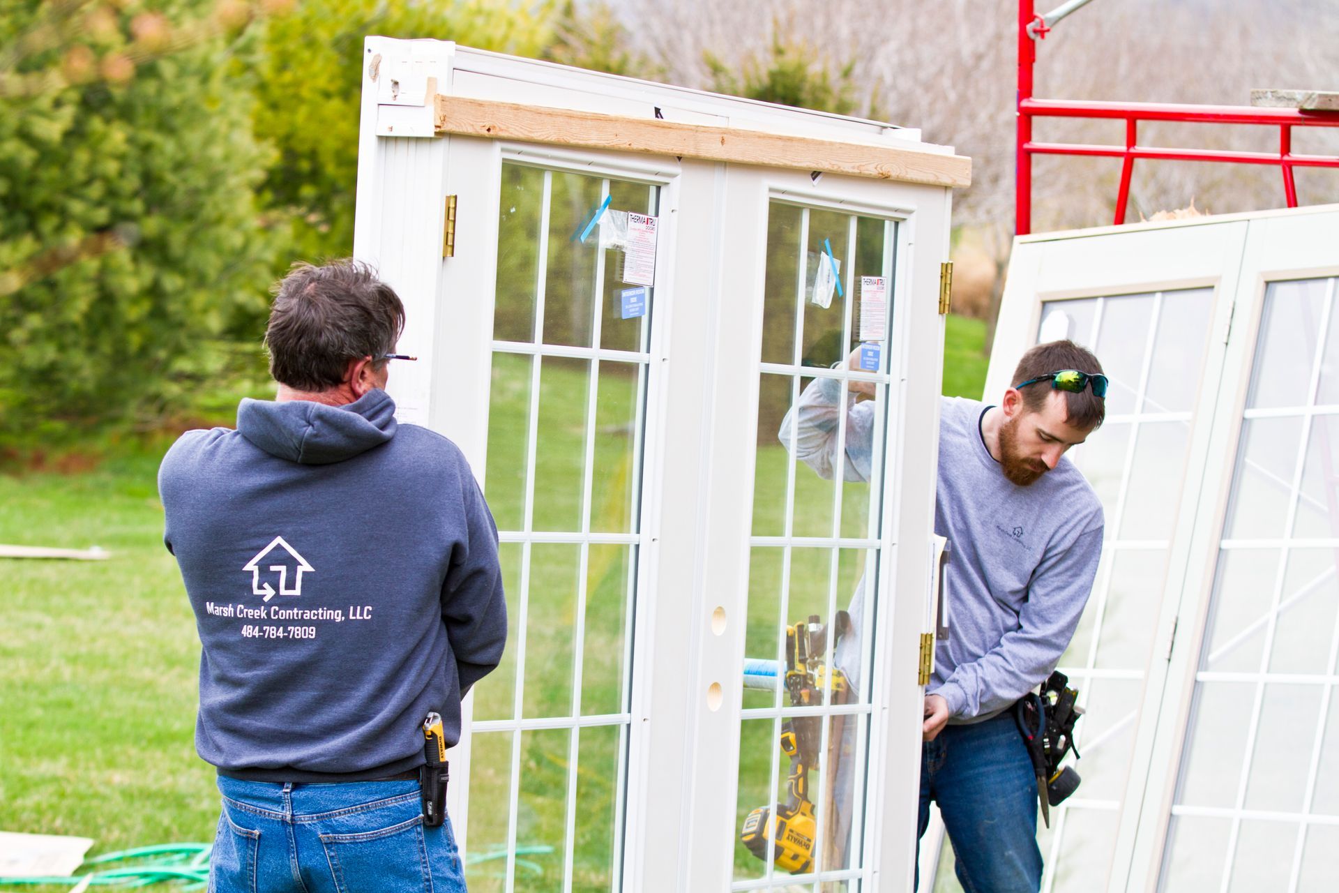 Two men carrying a white French door outside, likely installing it. One wears a hoodie with a logo.