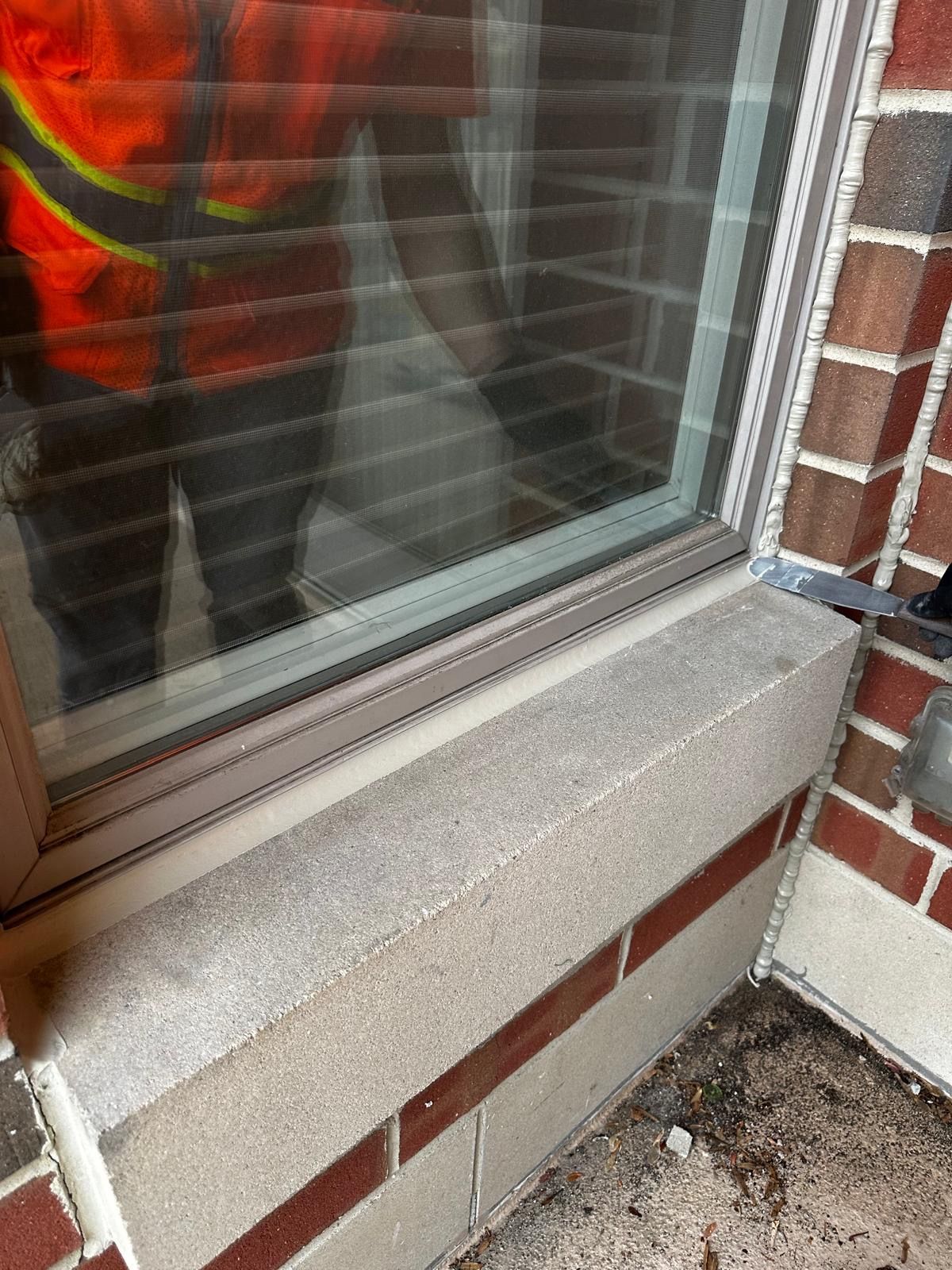 Window with gray sill, set in brick building. A person in an orange vest is visible through the window.