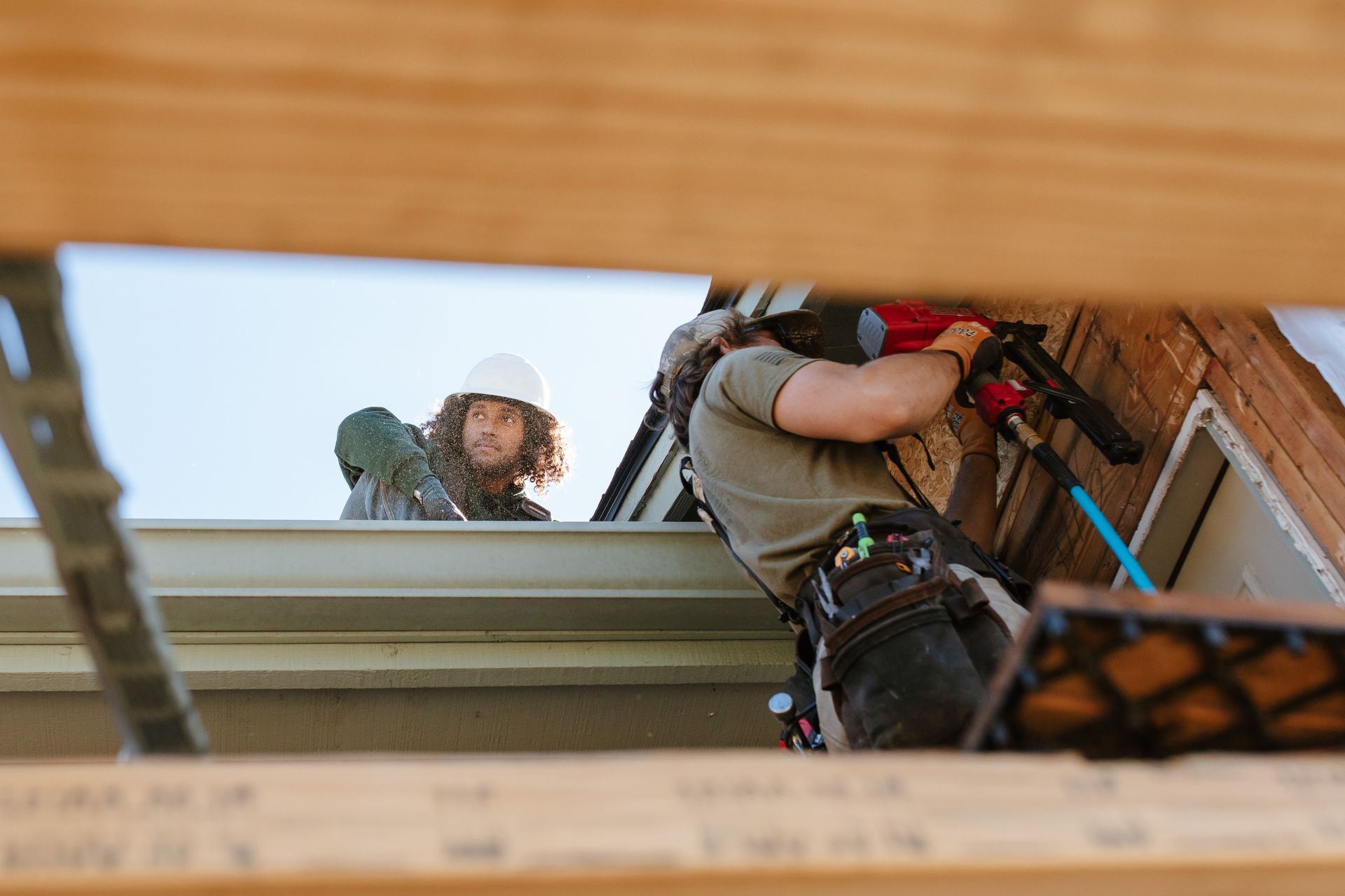 Two construction workers installing roofing, one using a nail gun.