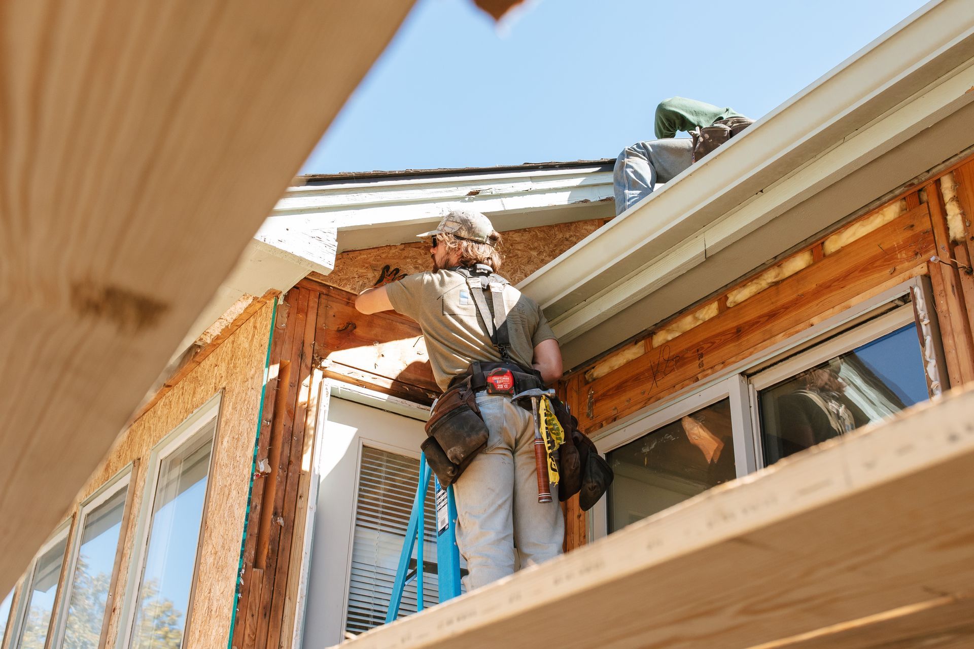 Construction worker on scaffolding, repairing a house. Sunlight, blue sky, tools, wood.