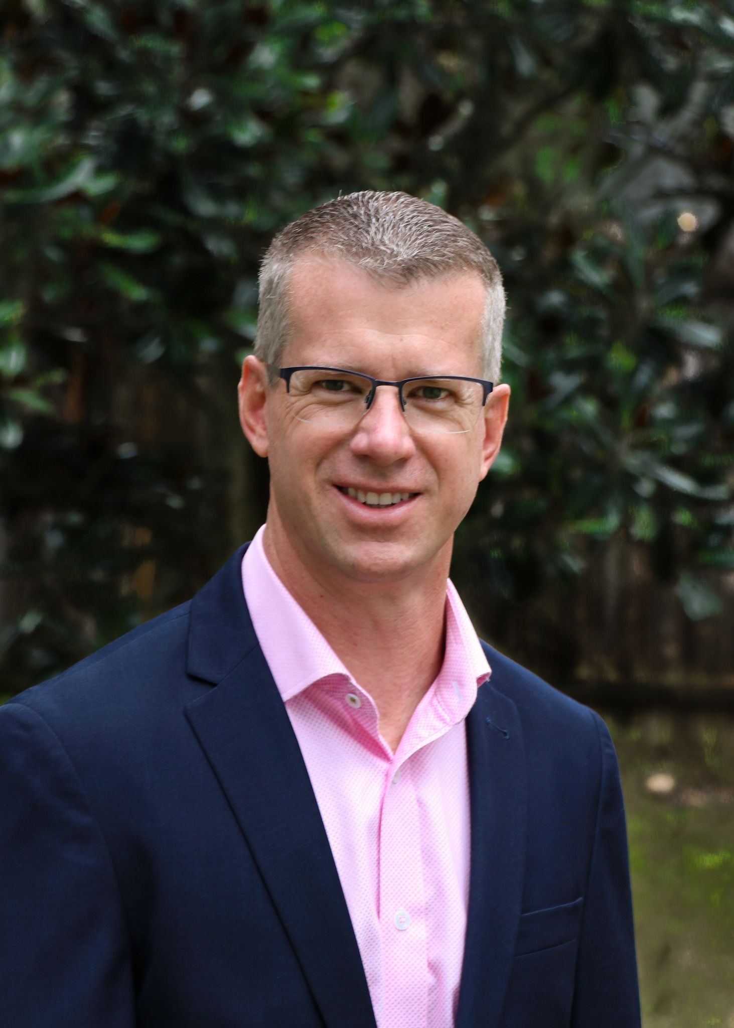 Man in glasses wearing a navy blazer and pink shirt, standing outdoors against a leafy background