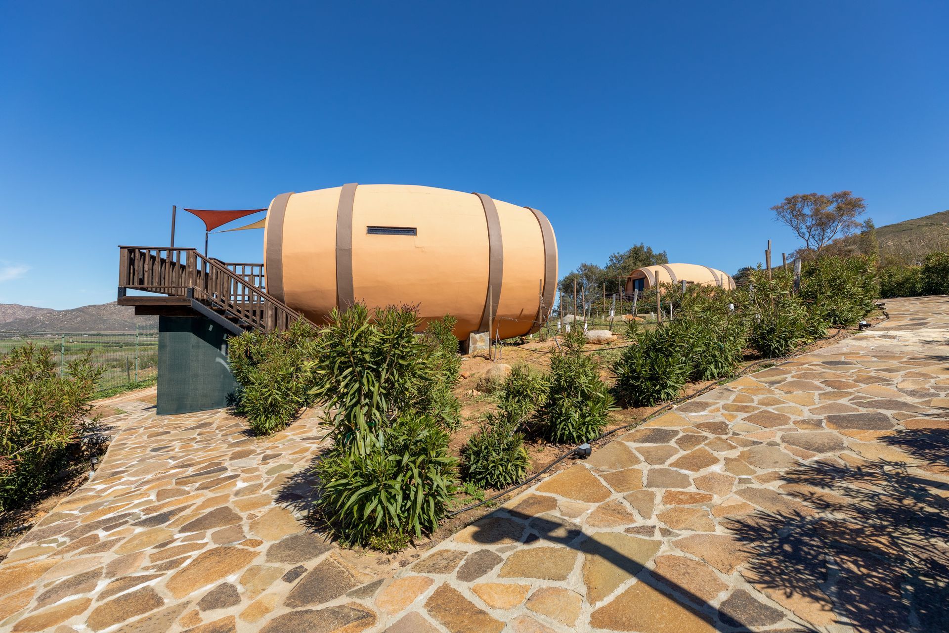 Pequeña casa cilíndrica de color tostado con terraza de madera en un camino de piedra bajo un cielo azul claro.