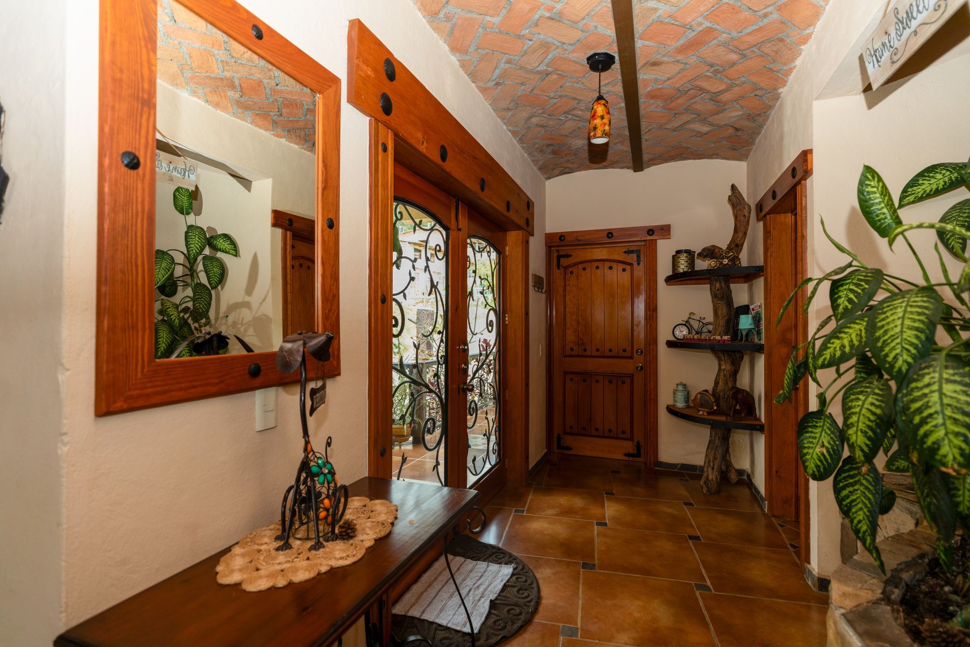 Pasillo estrecho con puertas de madera, techo decorado, espejo de pared y plantas en macetas sobre estantes y mesas.