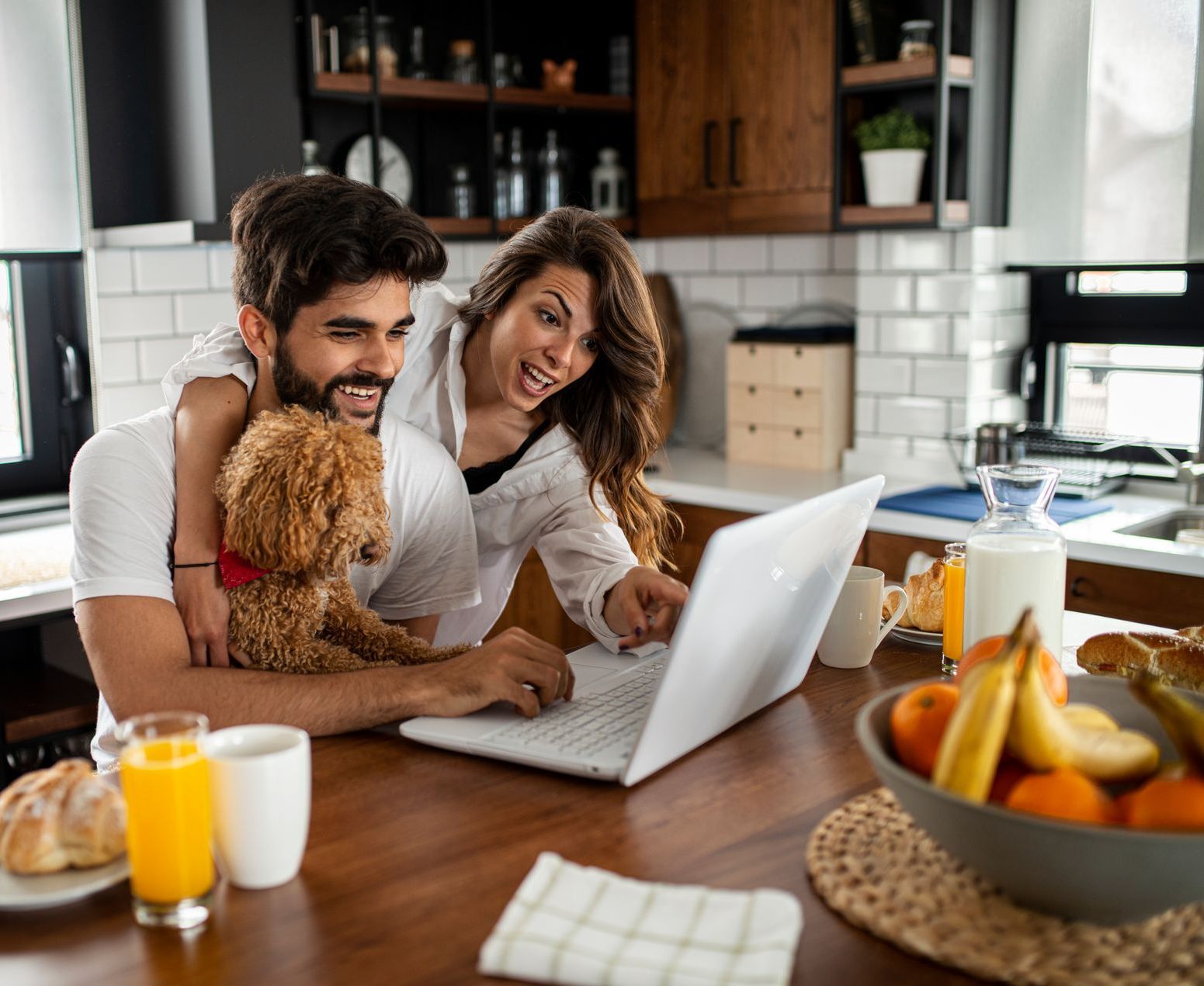 A man and woman are sitting at a table with a dog looking at a laptop.