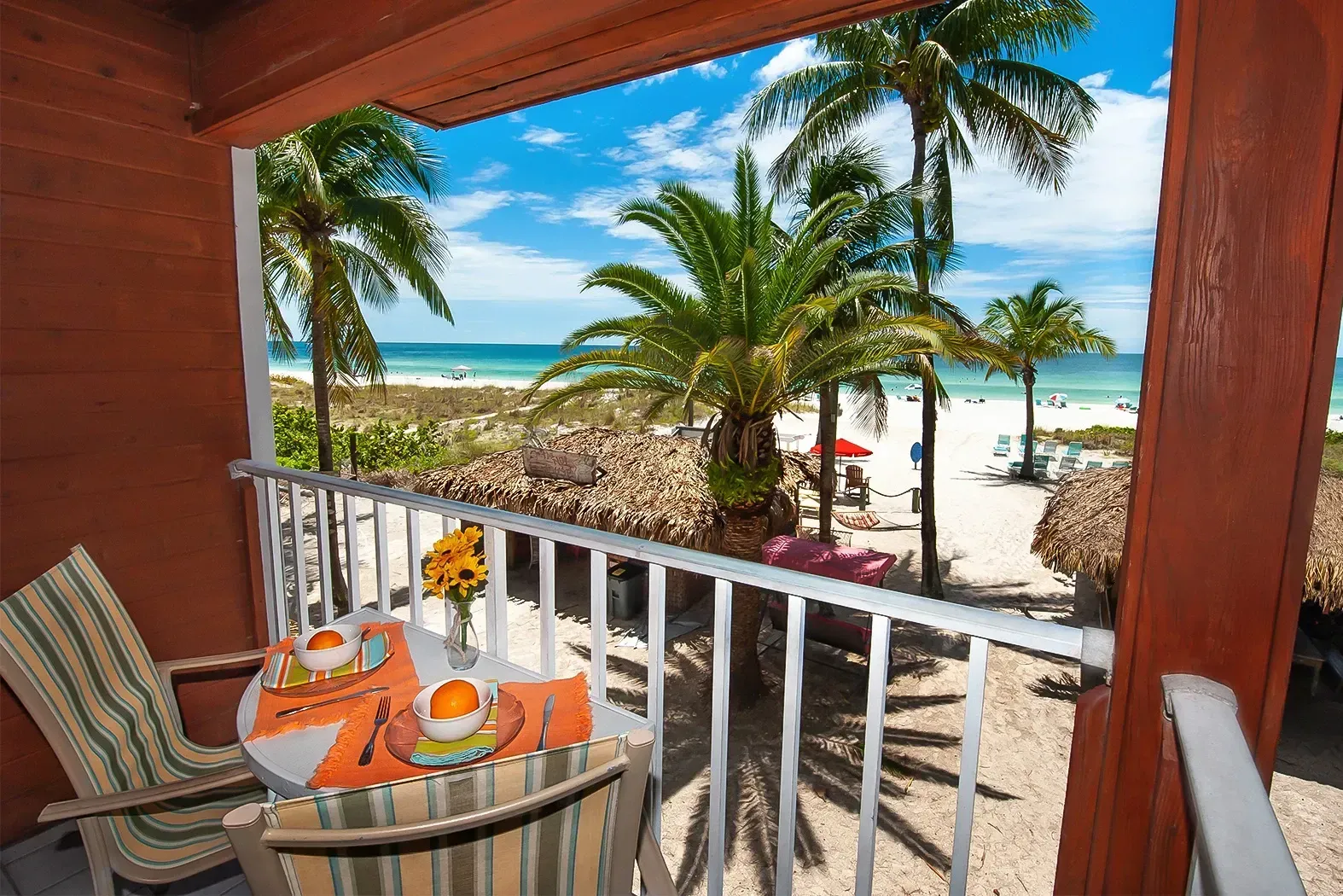 Balcony with table set for two overlooking a beach with palm trees and turquoise water.