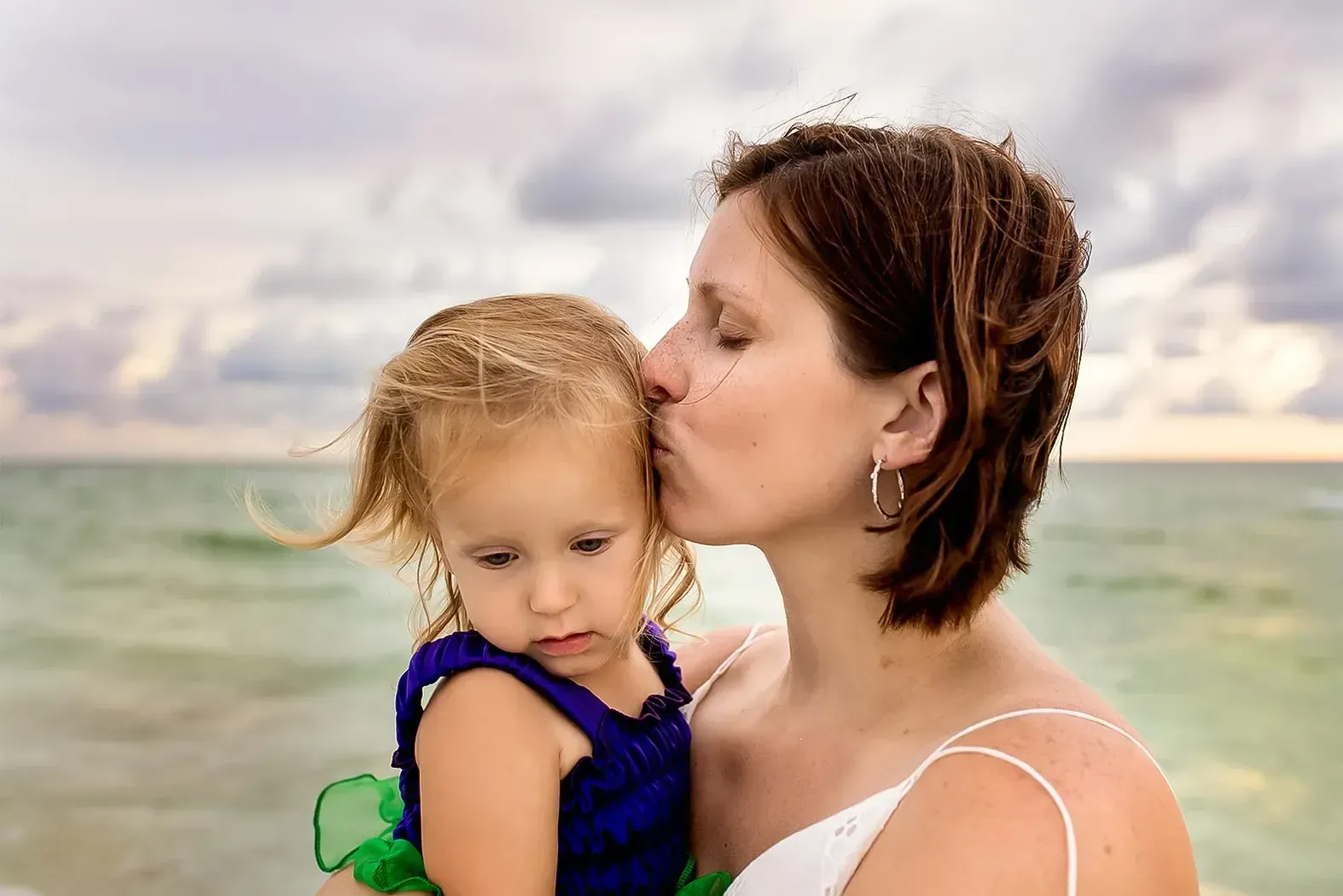 A woman is kissing her little girl on the cheek on the beach.