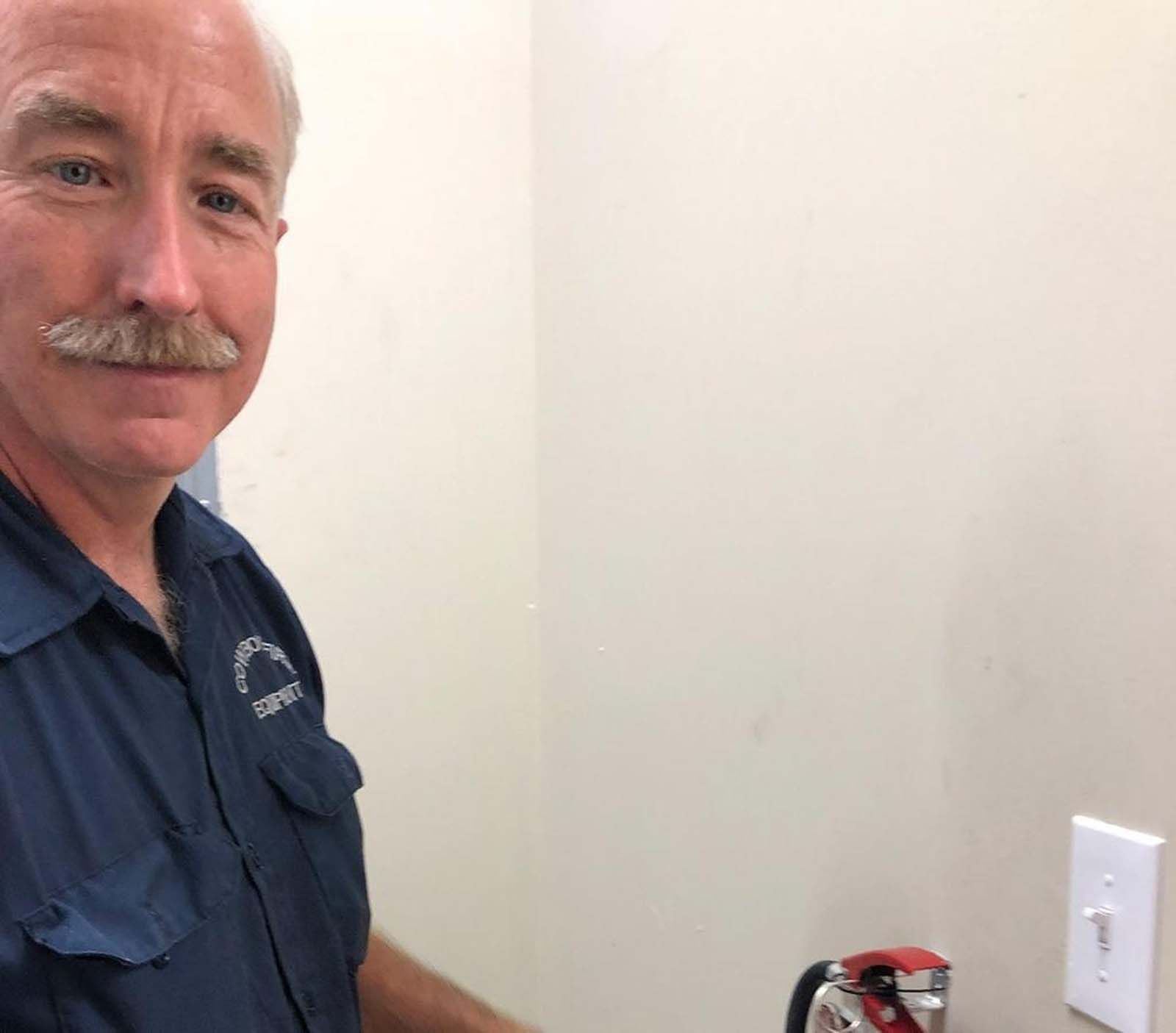Man with mustache in blue shirt stands near beige wall, light switch, and what looks like a fire extinguisher.