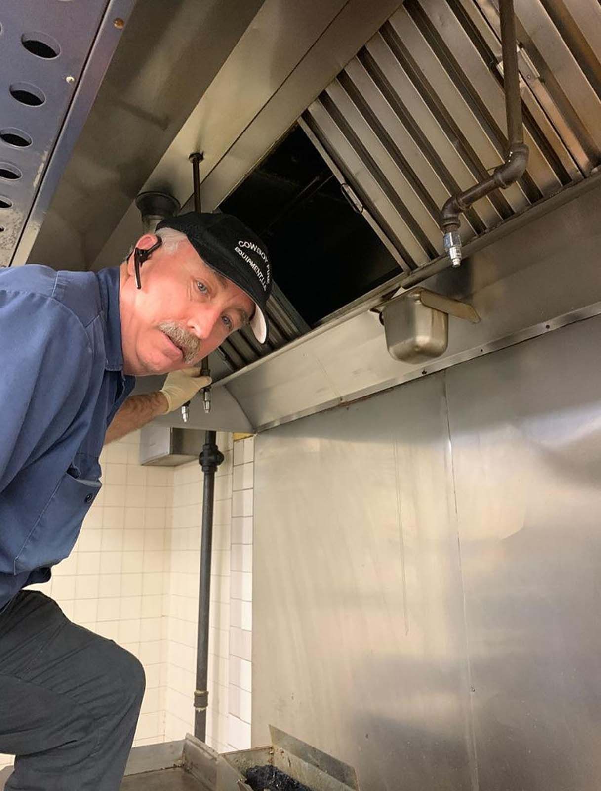 Man in cap inspecting a commercial kitchen hood. Stainless steel, tiled wall.