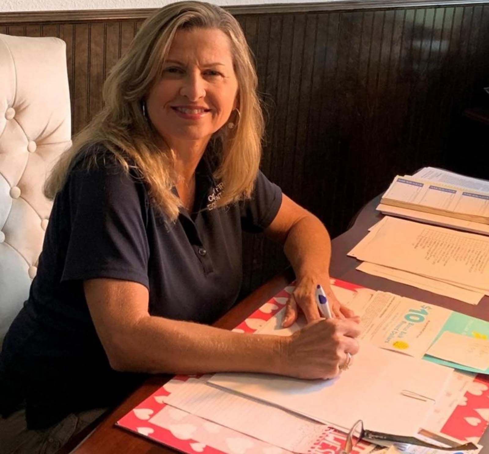Woman with blonde hair, smiling, seated at a desk, writing with a pen, papers and folders present.