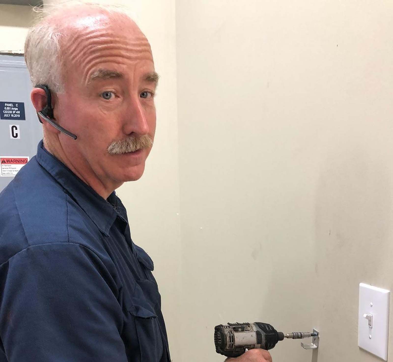 Man with mustache using drill on wall, wearing a headset, in a room with a light switch.