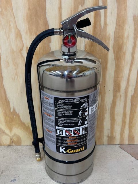 A stainless steel K-Guard fire extinguisher with a black hose and handle, mounted on a wood background.