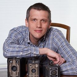 Man leaning on a table with three antique cameras, wearing a blue plaid shirt, smiling.